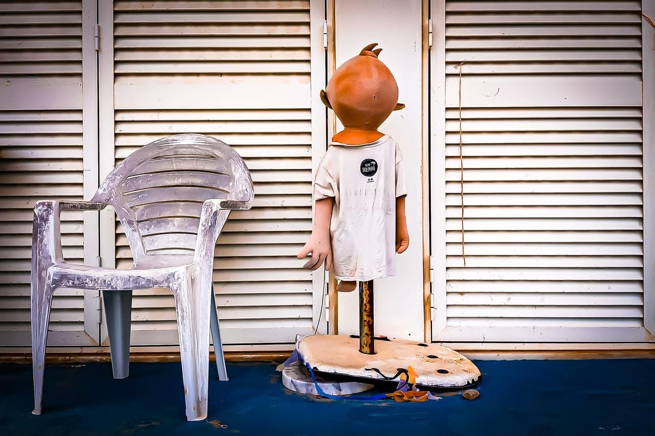 A weathered white plastic chair sits next to a mannequin with an oversized head, facing away. The mannequin wears a white shirt and is mounted on a stand with a cloth base on a blue floor. The background features white louvered doors.