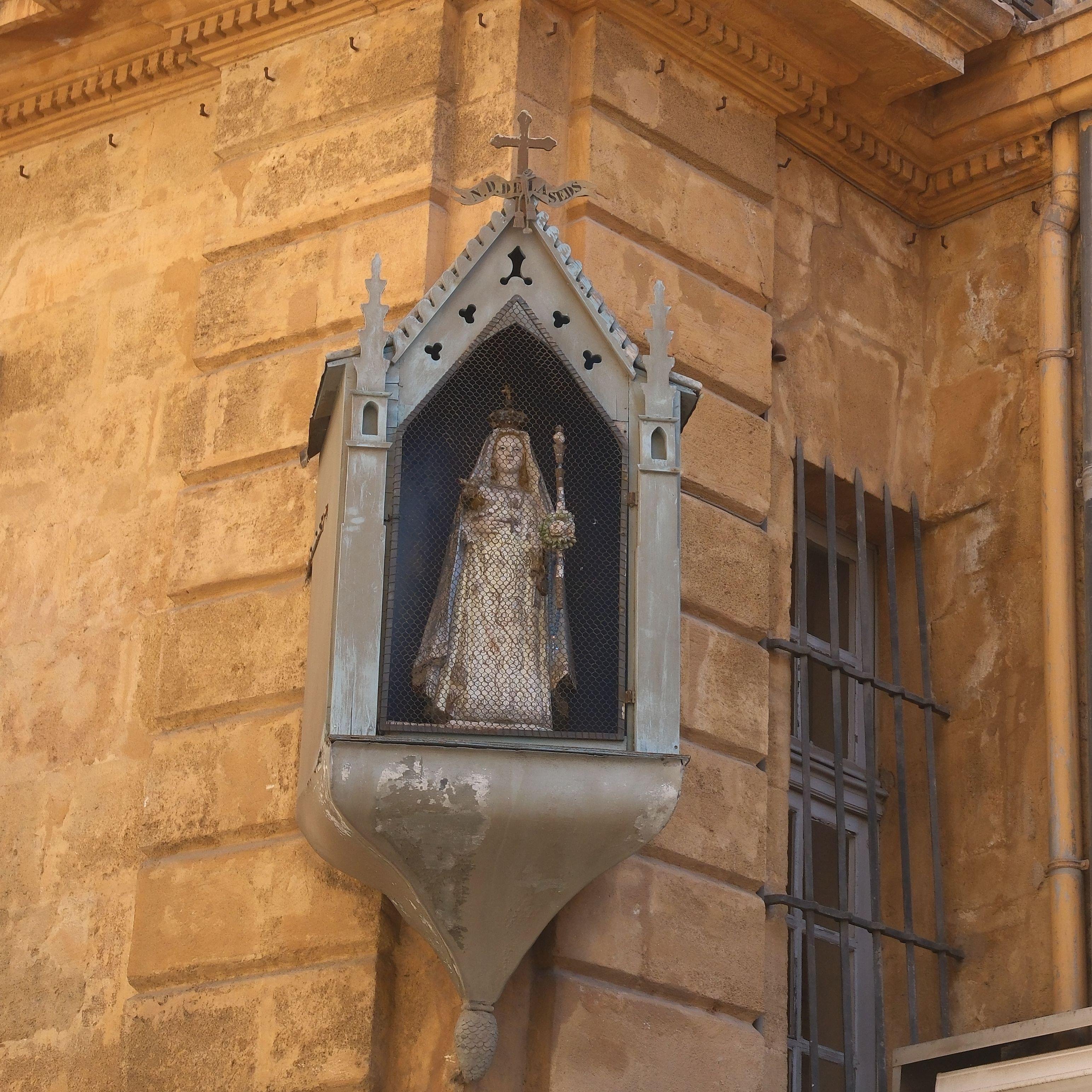 Photo carrée. Sur le coin d’un bâtiment couleur sable, une statuette représentant une sorte de prêtresse avec un sceptre et une couronne, à l’abri dans une mini chapelle en bois peint en gris