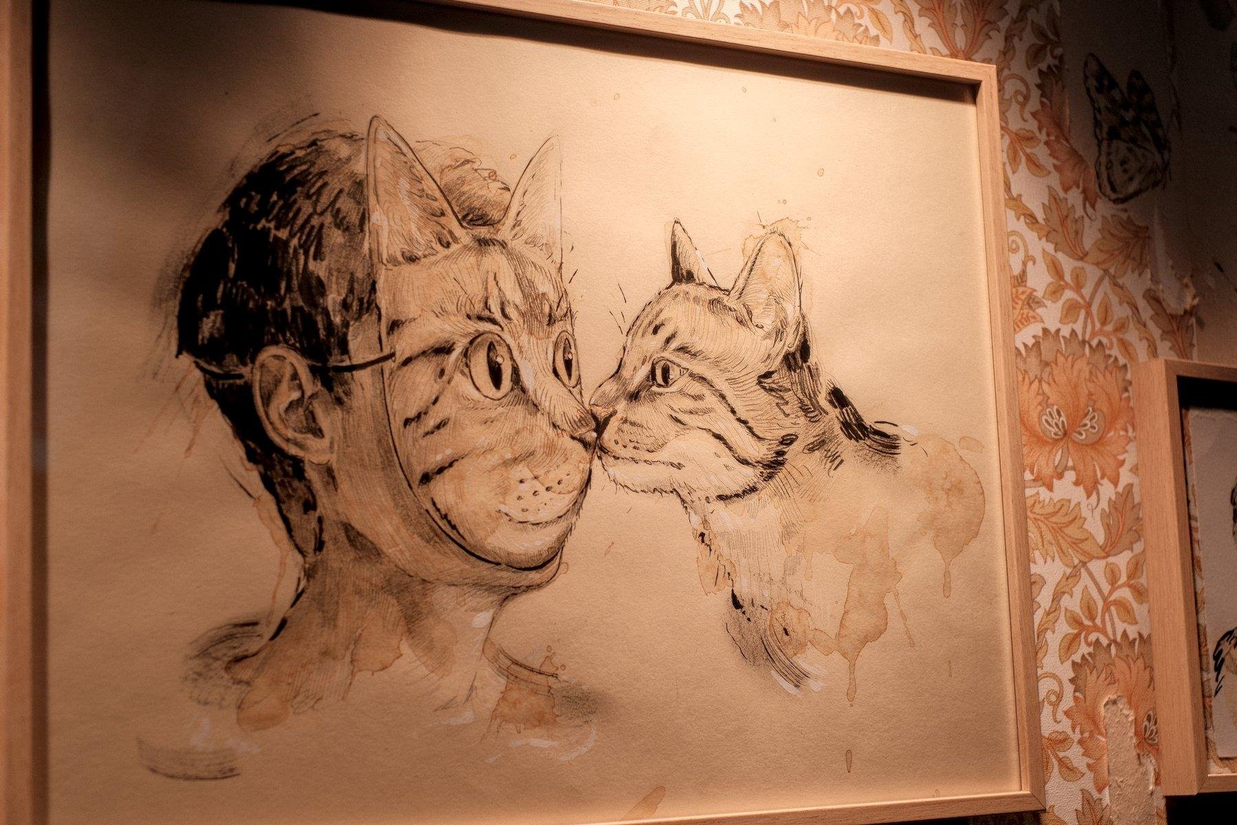 Photo horizontale. Une illustration encadrée et exposée représentant la tête d’une personne dont le visage est recouvert d’un masque représentant un chat. Il fait face à un vrai chat qui renifle le masque