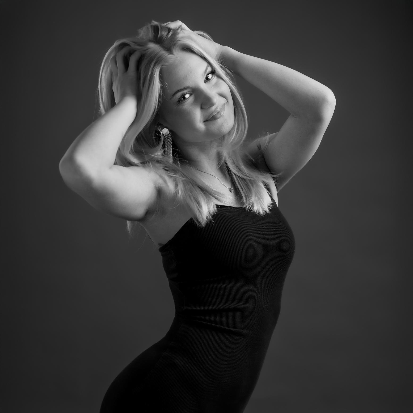 The image is a black and white portrait of a person with long hair, smiling and posing with their hands in their hair. They are wearing a sleeveless outfit. The background is plain, and the lighting highlights the subject.