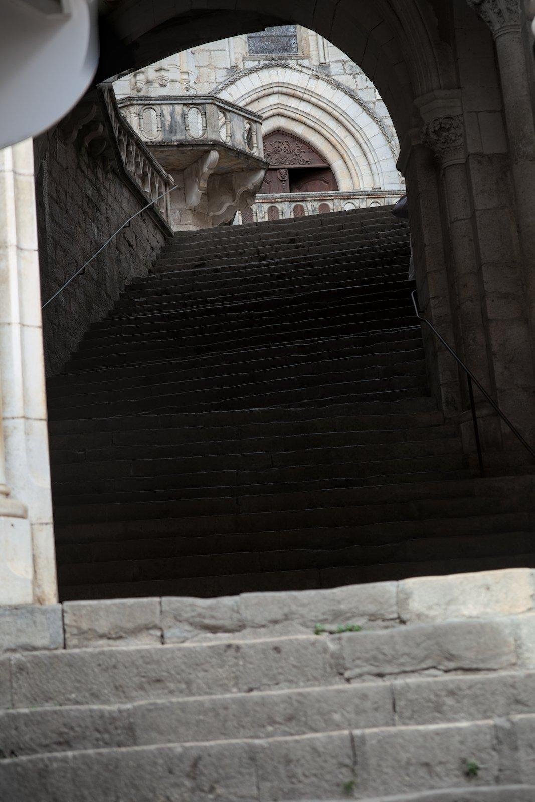 Des escaliers en pierre qui montent. On aperçoit une porte de chapelle au sommet