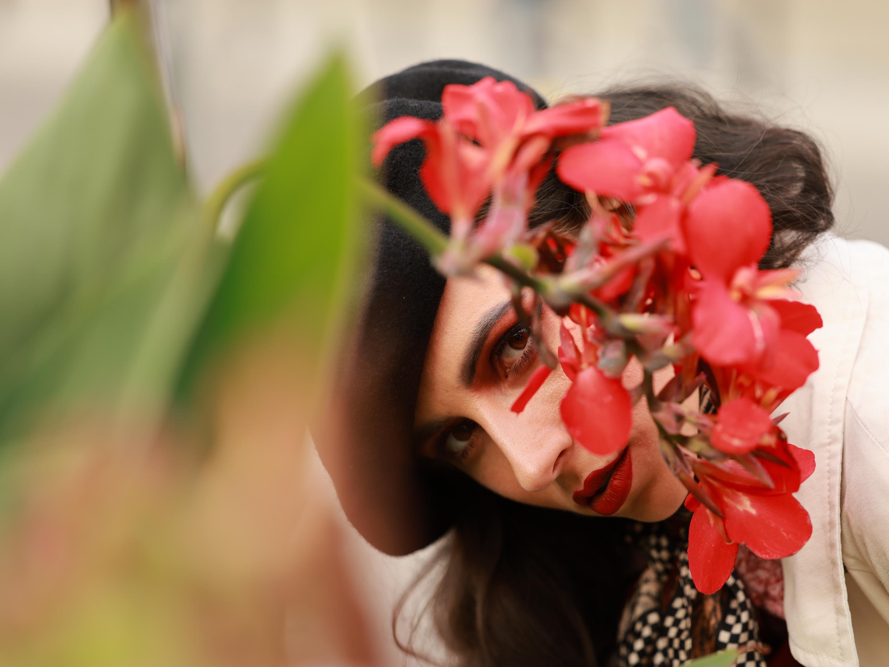Woman wearing a black beret and patterned scarf, partially obscured by red flowers in the foreground. She has long black hair and wears dark red lipstick, with a cream-colored garment visible. Focus on her eye peering through the flowers.