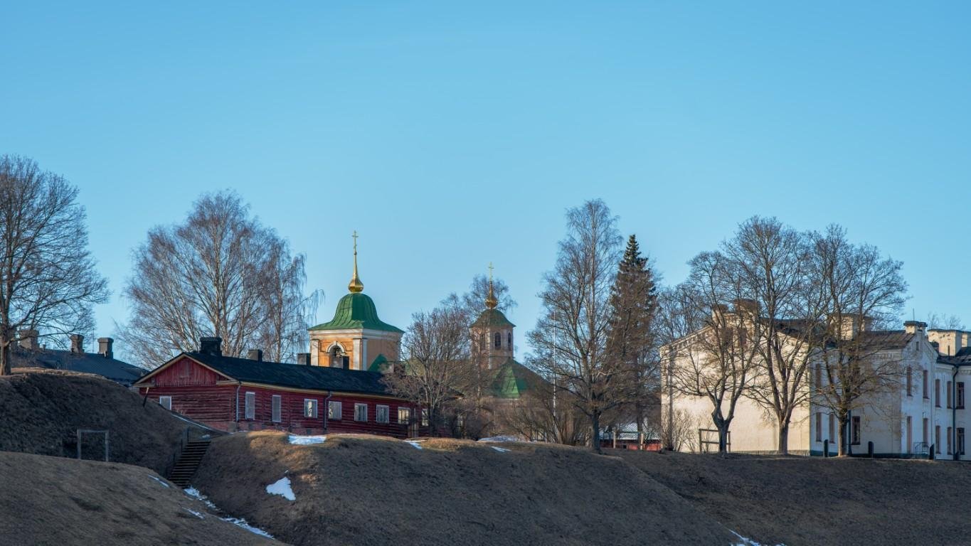 The image shows a scenic view with traditional buildings, including a wooden structure and a church with green and golden domes. Bare trees are visible, suggesting it's either late winter or early spring. The sky is clear and blue, adding to the serene atmosphere of the scene.