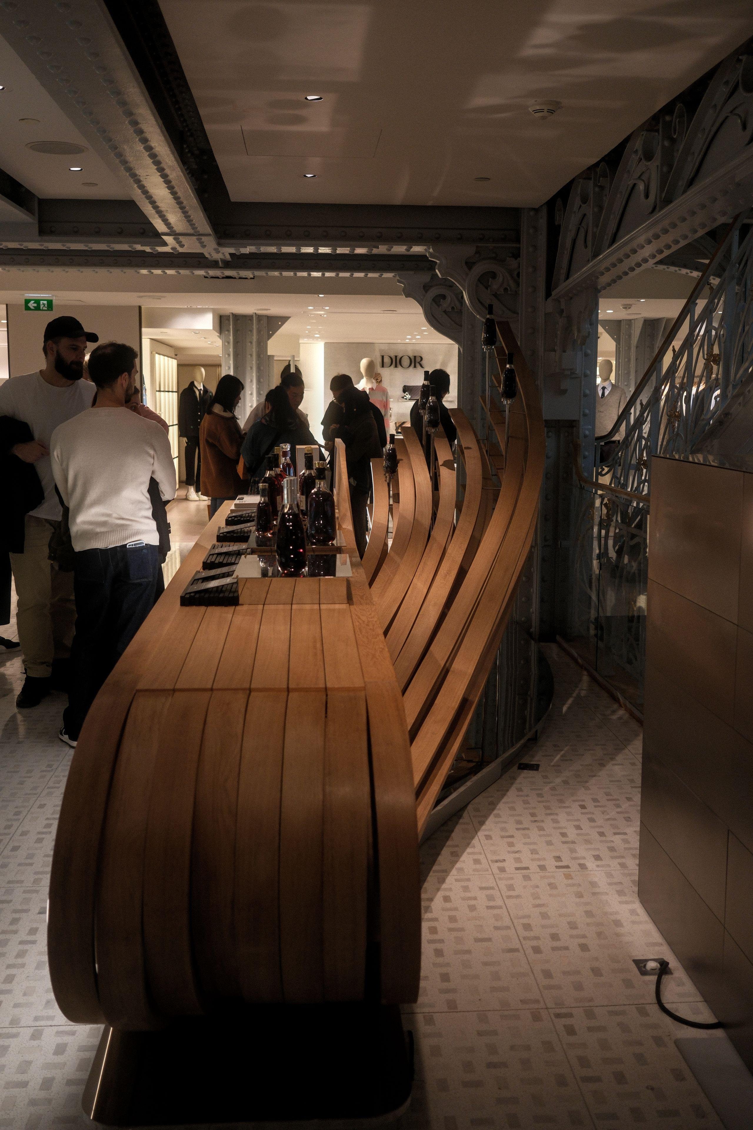 Photo verticale. Un présentoir fait de lattes de bois, assez pour faire une surface plane où sont posées des bouteille de ce qui semble être du vin. Derrière, d’autres lattes « sortent » du comptoir en se tordant vers le haut pour former une décoration moderne.