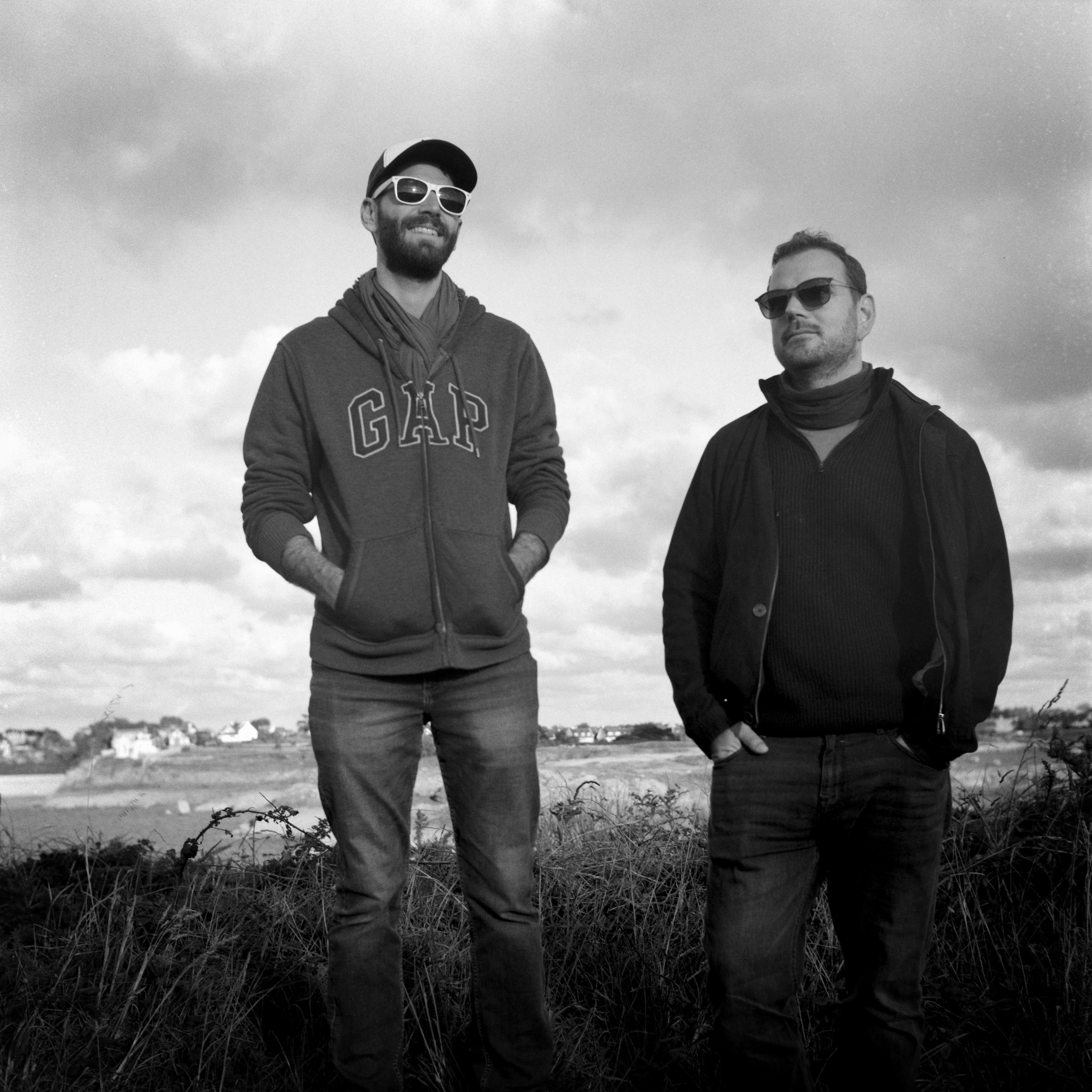 This is a black and white image of two men standing outdoors in a natural setting. They are both wearing sunglasses and casual clothing, including jackets. One man is wearing a cap and a hoodie with the word "GAP" on it. The background shows some clouds in the sky and a coastline.
