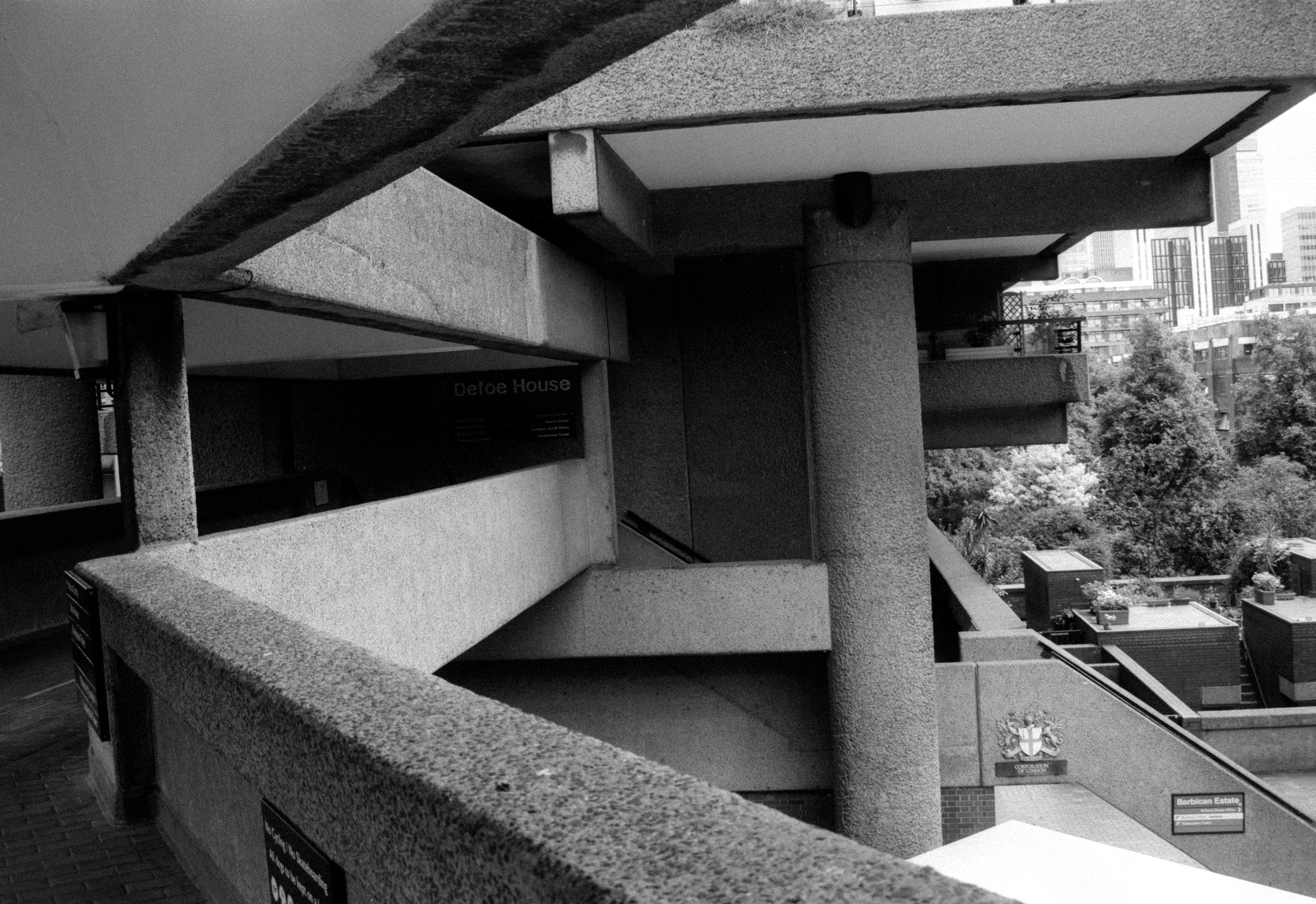 Taken from the walkway by Defoe House in the Barbican Estate in London. Though the photograph is in black and white, everything is constructed of grey concrete, so it would not make too much difference. The camera is pointing towards Defoe House itself, its name just visible in the shadows on the left. There is a barrier at the bottom of the frame designed to keep pedestrians safe. There is a large support pillar visible near the middle of the frame, with a balcony right next to it. To its left is the staircase that takes you to the ground. On that staircase nearer the ground is the Corporation of London logo, and a Barbican Estate information sign.