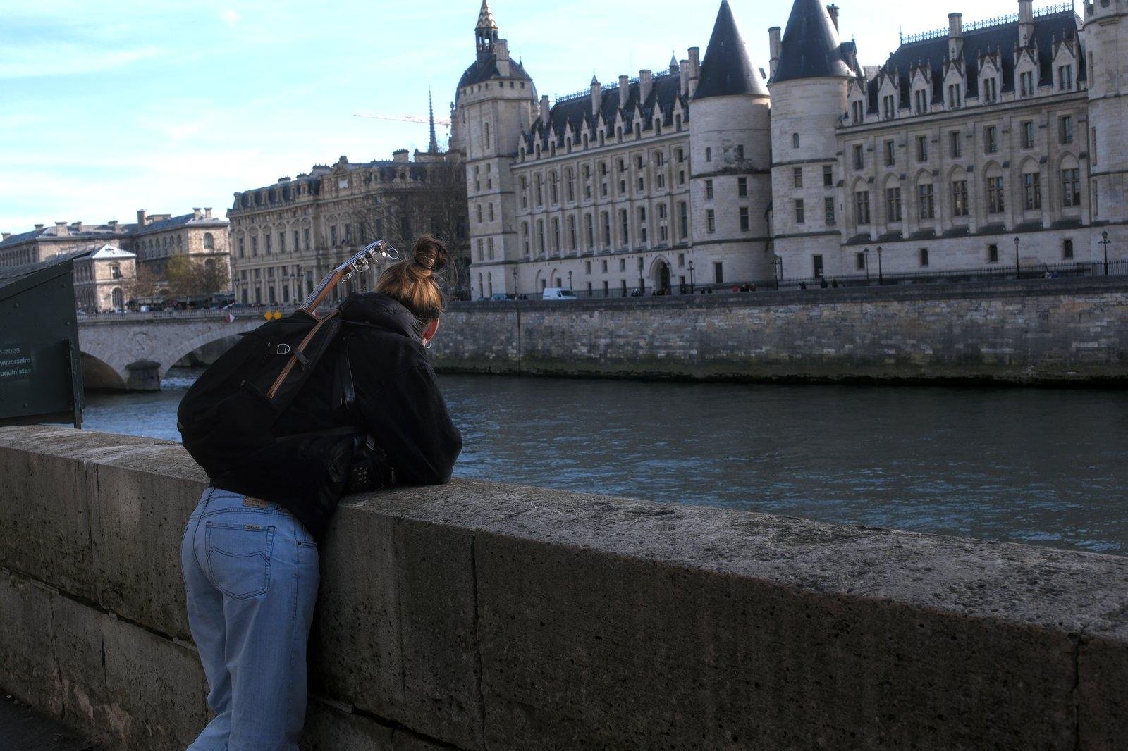 Photo horizontale. Une jeune femme chatain coiffée en chignon rapide, habillée en jeans et manteau court noir, avec un sac à dos noir dont dépasse le manche d’un ukulele, est accoudée à une rambarde en pierre au dessus des quais de Seine parisien et regarde en contre bas. On voit la Seine au second plan et l’autre rive en arrière plan