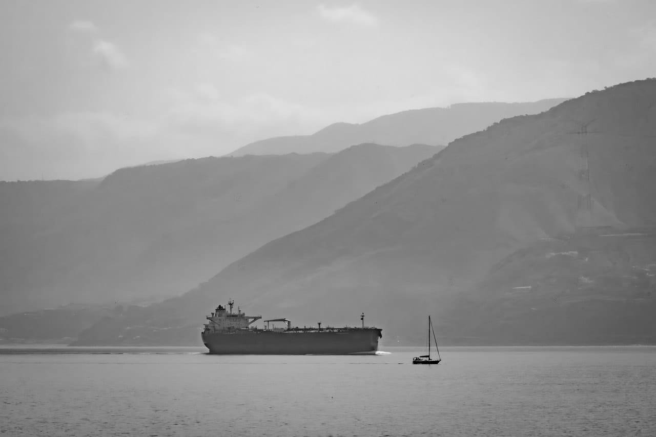 Black-and-white seascape with a large cargo ship on calm water near the shore, a small sailboat in the foreground, and layered mountain ridges in the hazy background under a cloudy sky.