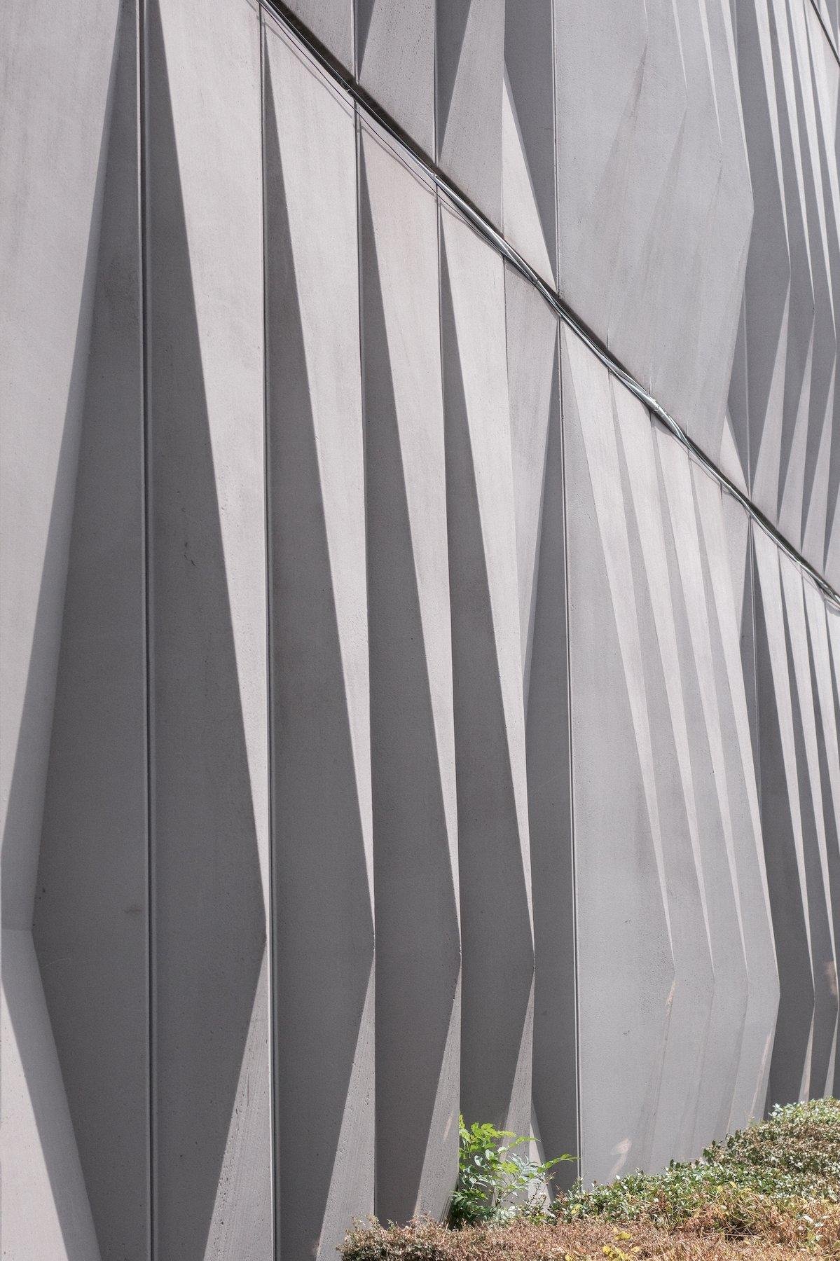 Photo verticale. Un mur fait de formes angulaires qui se répètent, donnant 4 teintes de gris selon la face