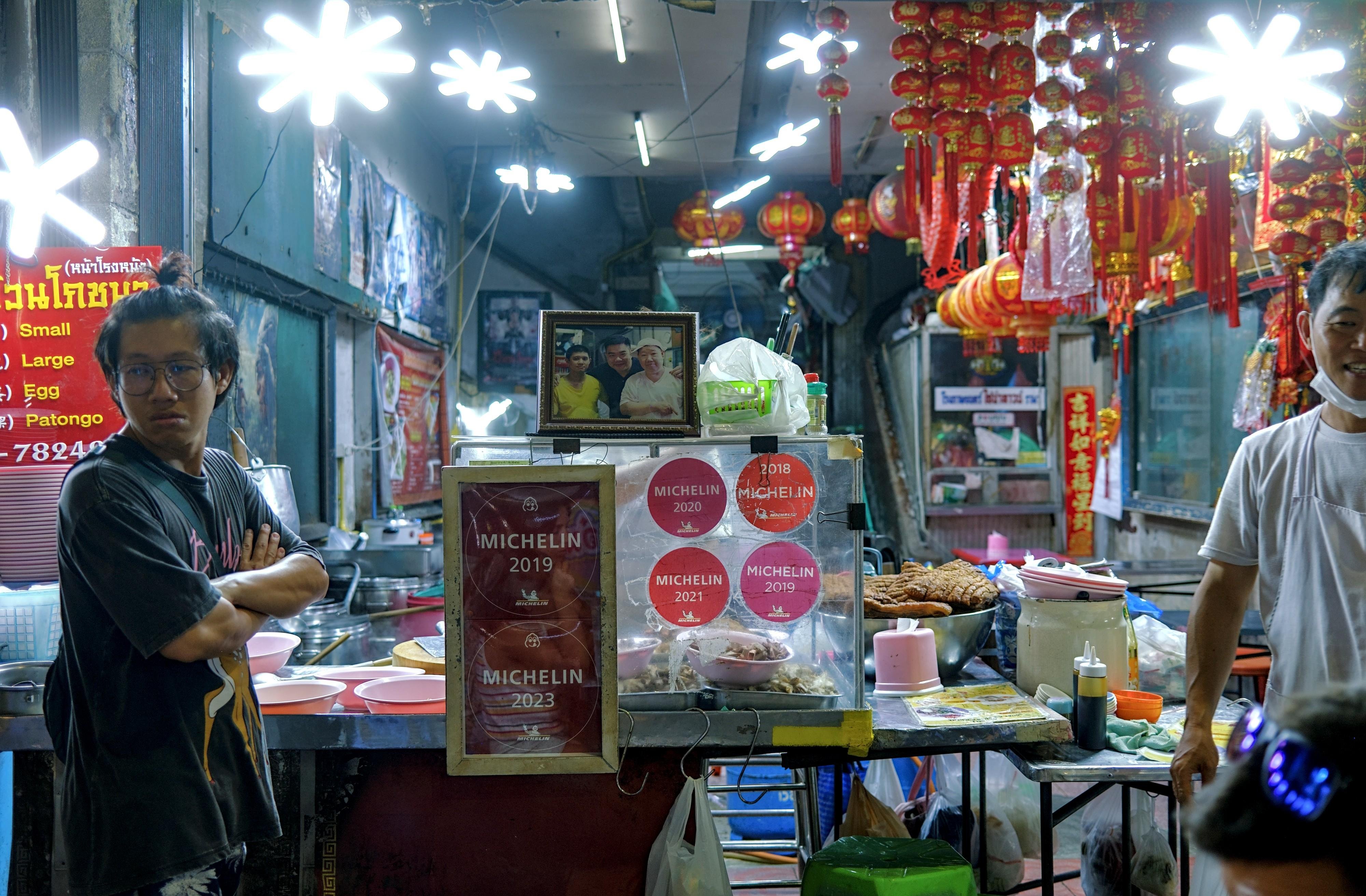 A man looks unconvinced, off camera. Behind him, a food stall with a bunch of Michelin stickers.