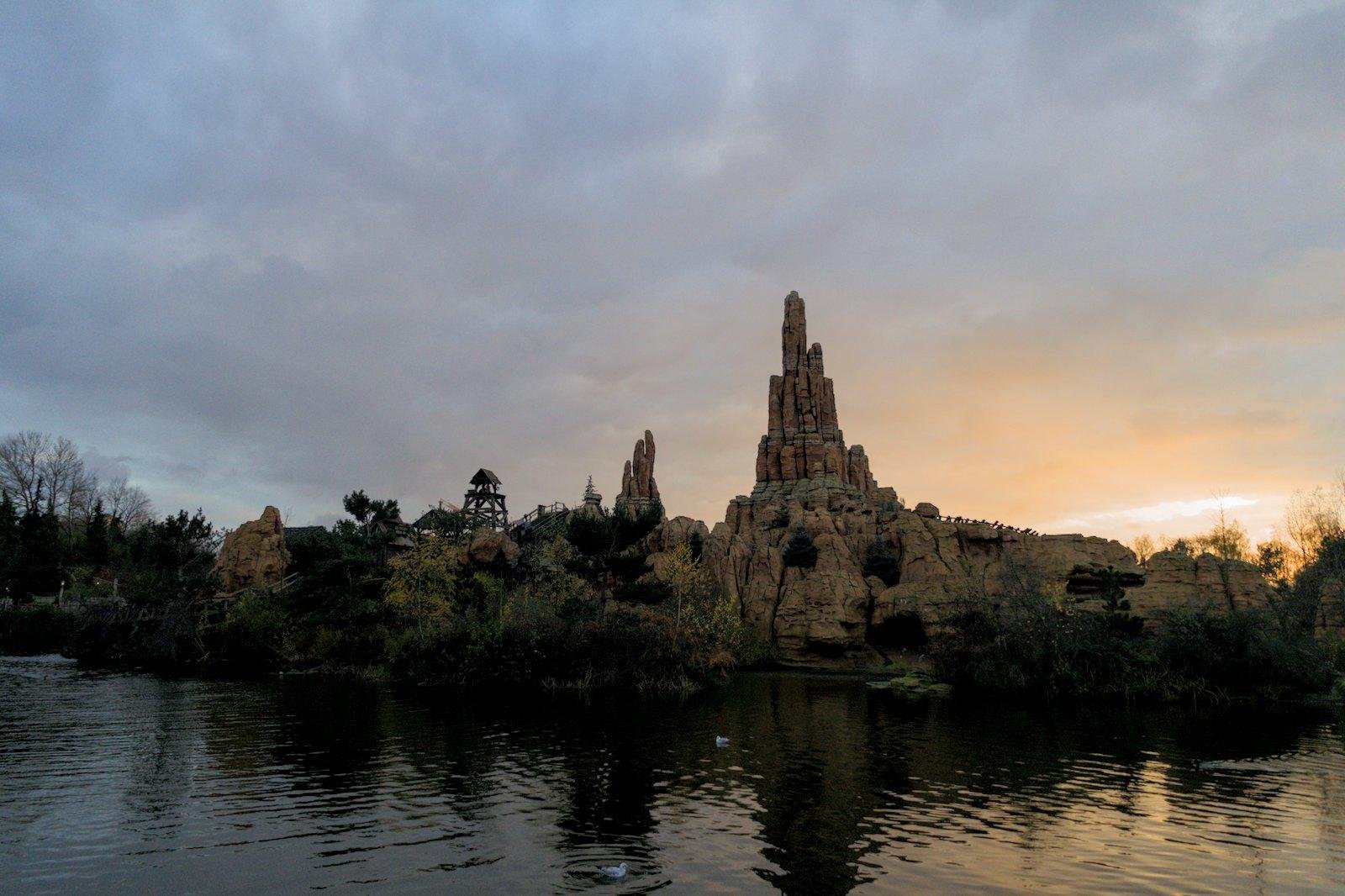 Photo format paysage de la mine à Disneyland Paris. Le premier plan est plein d’eau, le second est la montagne de la la mine avec son grand pic caractéristique et des arbres à ses pieds et le ciel affiche un soleil couchant, faisant un dégradé orange vers le bleu pale