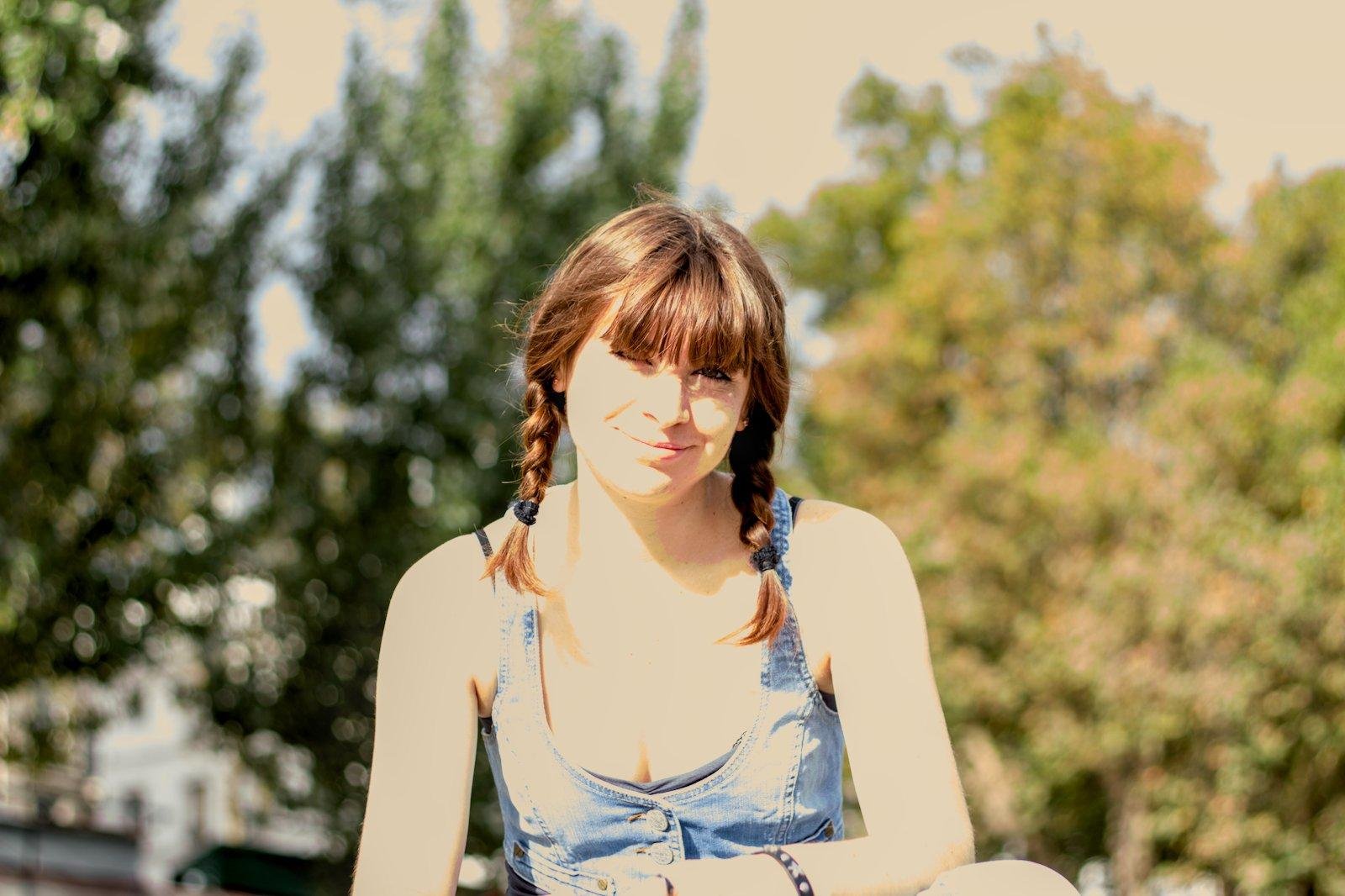 Photo format paysage montrant une jeune femme brune avec des nattes, regardant la caméra en souriant. Elle porte un haut en denim, le fond est constitué d’arbres flous. La photo est surexposée donnant une impression de forte chaleur