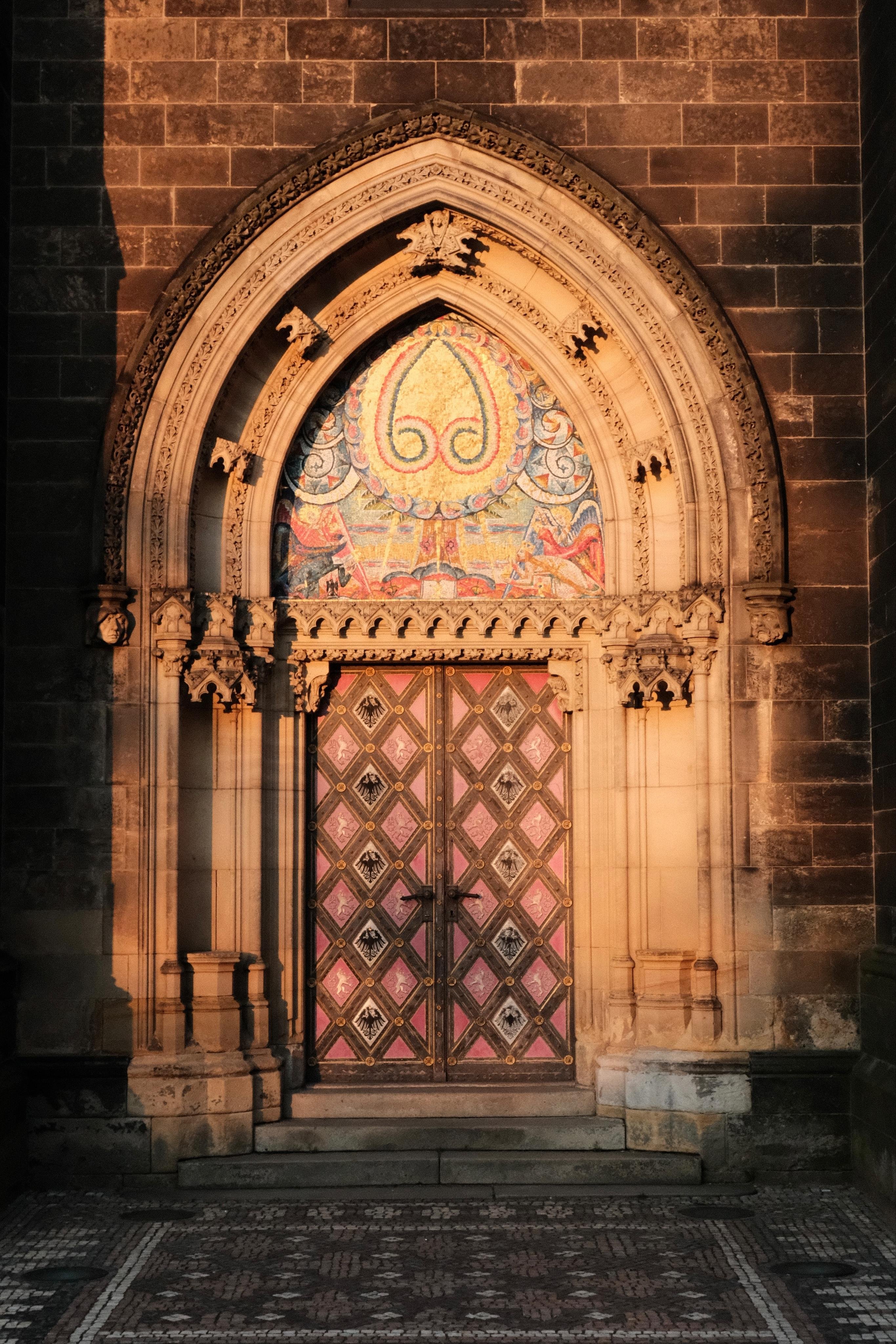 Gothic-style stone doorway with a pointed arch and carved details, featuring double wooden doors with a pink and gold diamond pattern and a colorful mosaic panel above, set in a stone wall with steps leading up from a patterned stone walkway. The door is selectively highlighted by a patch of warm sunlight, while the surrounding arch and wall is in darker shadow.