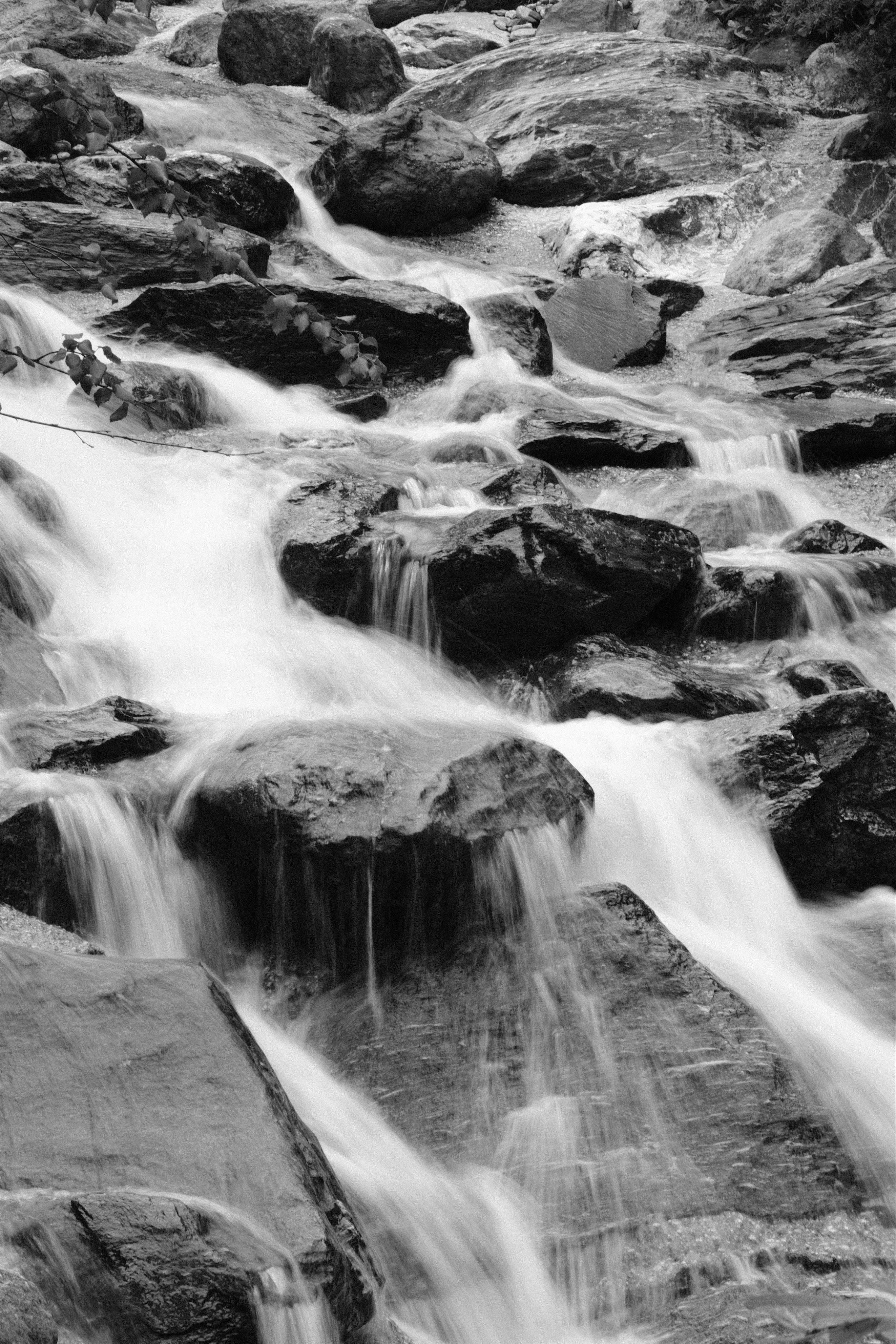 A black and white, longer exposure of a rocky mountain riverbed. The water energetically flows around larger rocks and boulders down the steep slope. Due to the longer exposure, the flow of water is visible, rather than sharp splashes.