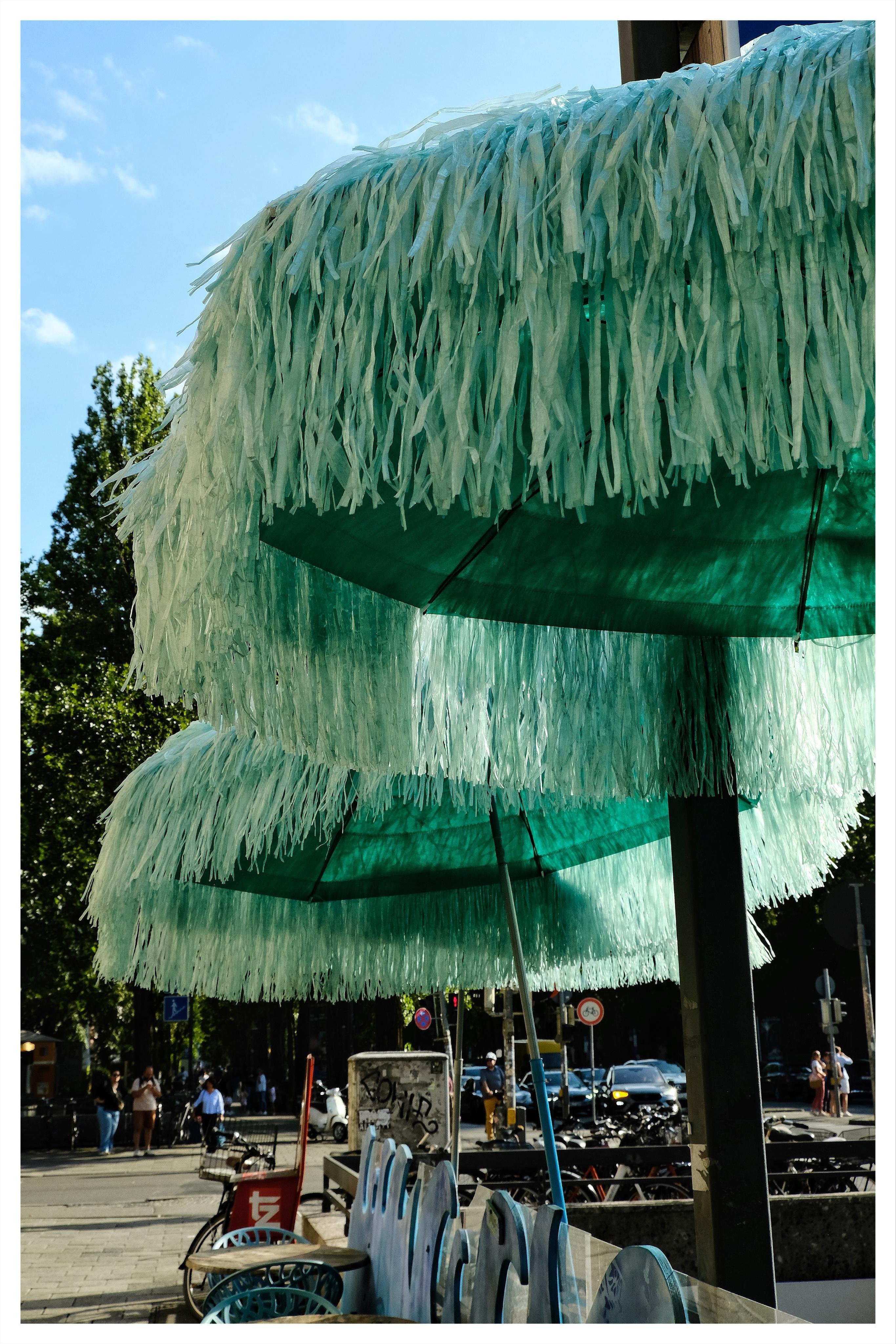 Two teal patio umbrellas with long shredded fringe stacked one above the other on a city sidewalk, with pedestrians, bicycles, cars, and trees in the background under a blue sky.