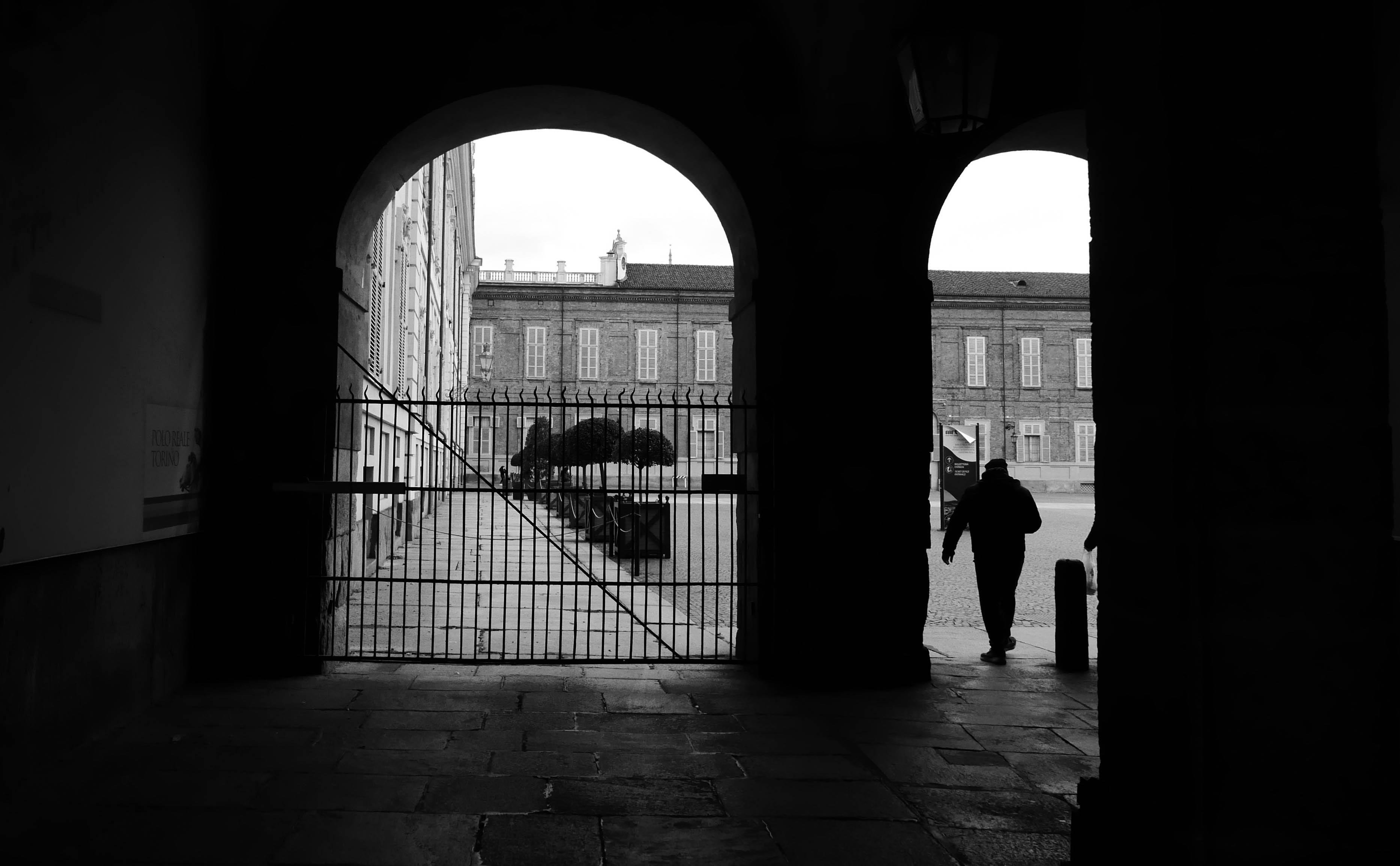 The image is a black and white photograph showing an archway leading to a courtyard. There is a gate in the foreground, and through it, you can see a person silhouetted against the light, walking towards the courtyard area. The background features a large building with uniformly spaced windows.
