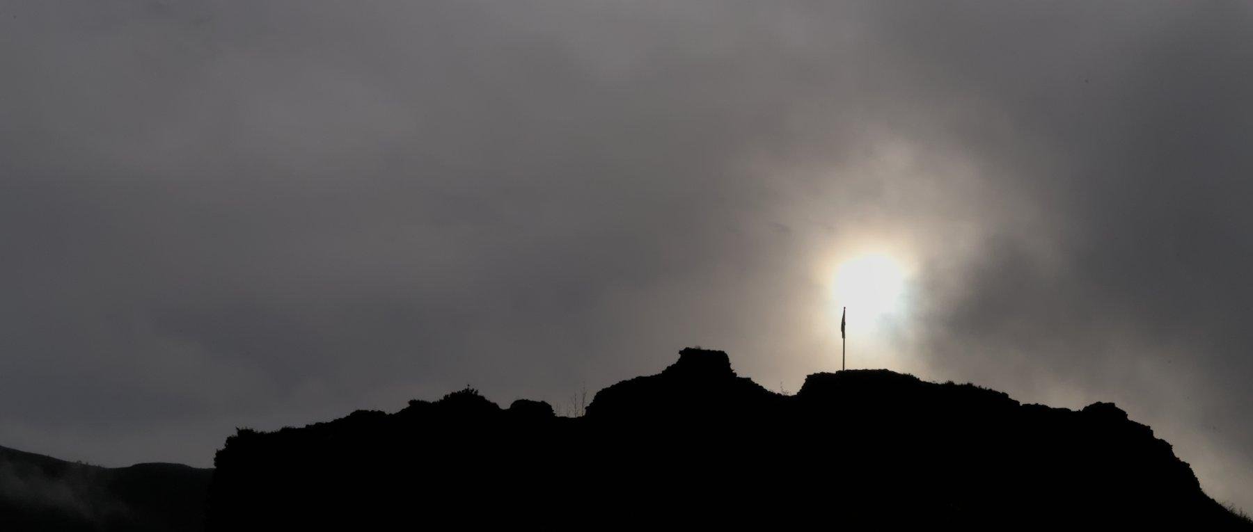 Photo horizontale. Contre jour du sommet d’une montagne sur fond de ciel gris. Le soleil perce derrière les nuages et la silhouette d’un pic de drapeau se dessine sur le cercle solaire