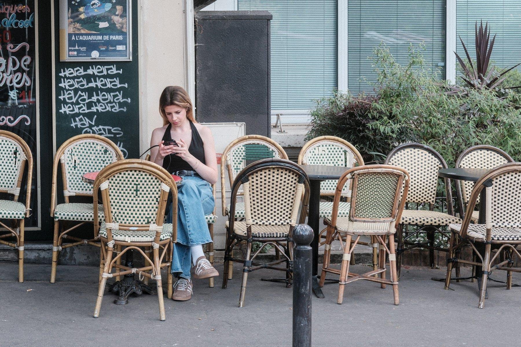 Photo horizontale. Une jeune femme aux cheveux long châtain aux reflet blonds, en jean basket et haut sans manche noir qui s’attache dans la nuque, assise seule à une terrasse de café, en train de consulter son téléphone