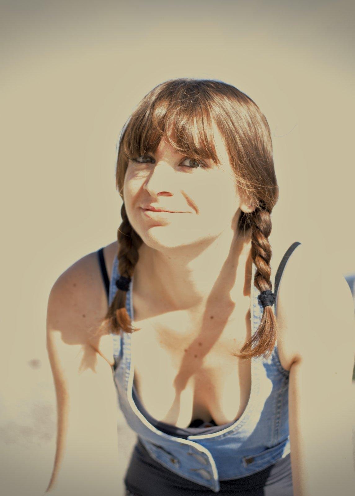 Photo format portrait au ton sépia, d’une jeune femme au cheveux brun coiffés en 2 nattes qui pendent devant elle. Elle est habillée d’un débardeur noir et par dessus, un gilet en denim. Elle regarde la caméra avec un air amusé