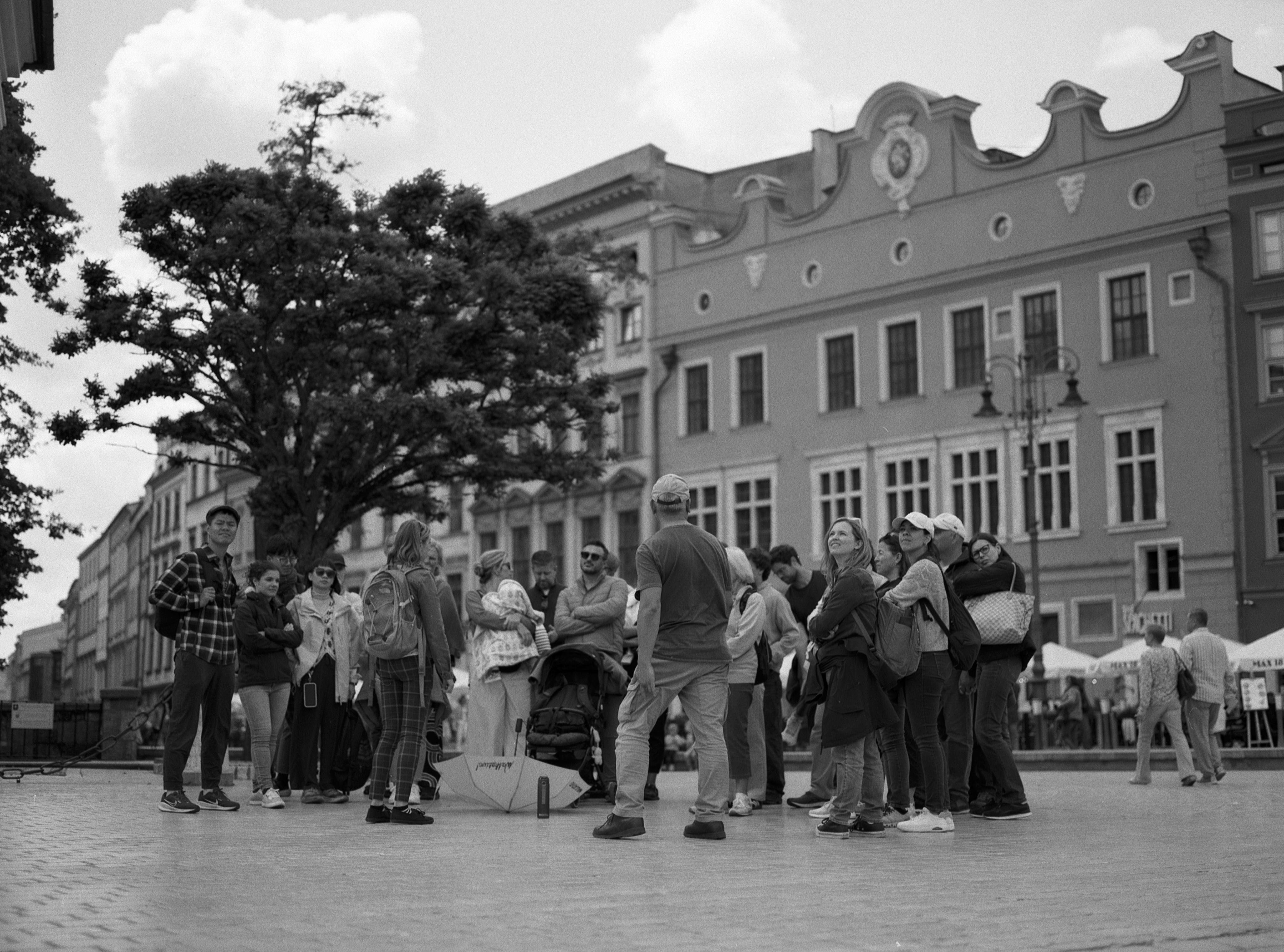 Group of people standing in a cobblestone street, some looking upwards and others engaging in conversation. A large tree and ornate buildings with detailed facades are in the background. Overhead, the sky is partly cloudy. Image is in black and white.