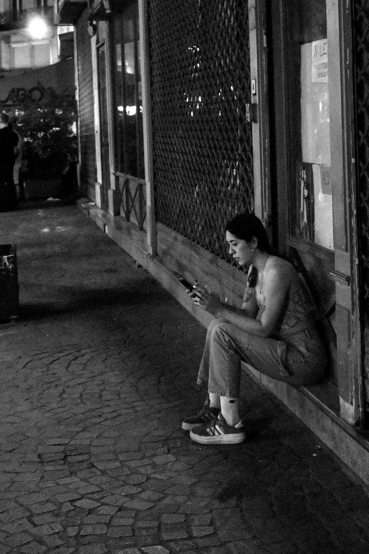 Photo verticale en noir et blanc de nuit. Une jeune femme en basket, jean et débardeur, assise sur une marche de magasin fermé, en train de consulter son téléphone