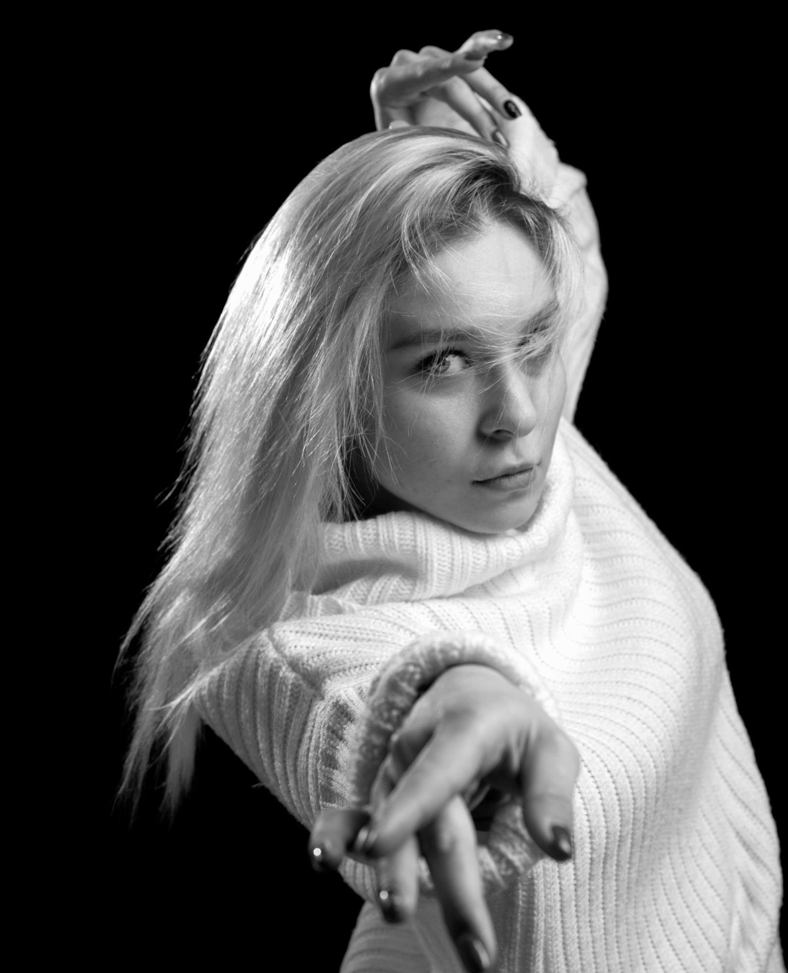 A woman with long blonde hair wearing a white knitted sweater extends her left hand towards the camera. Black fingernail polish is visible on her fingernails. Her right hand is placed behind her head. The background is completely black, creating a strong contrast with her light-colored clothing and hair.