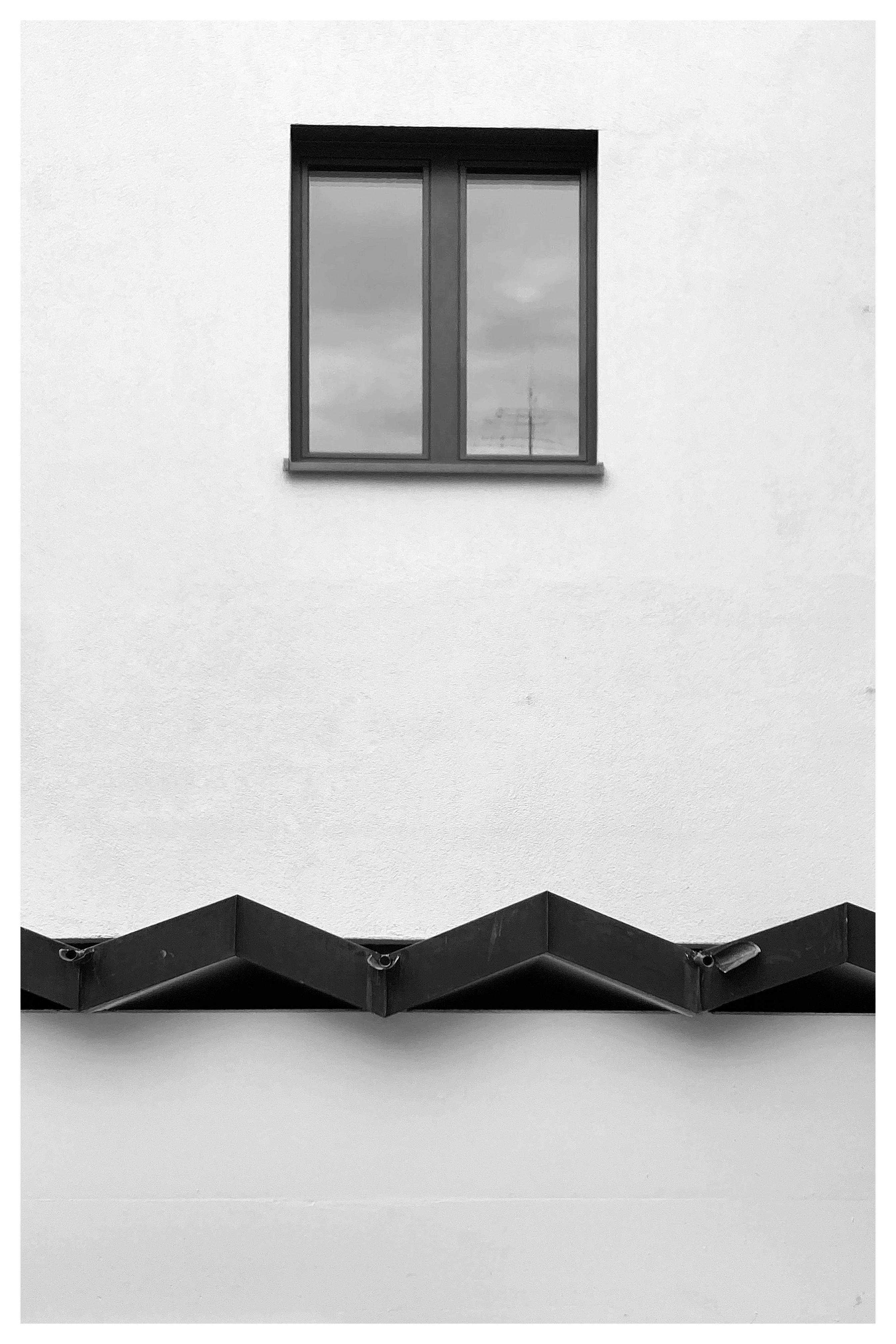 Black-and-white photo of a plain light-colored building facade with a centered two-pane window near the top and a dark zigzag roof edge across the lower portion.