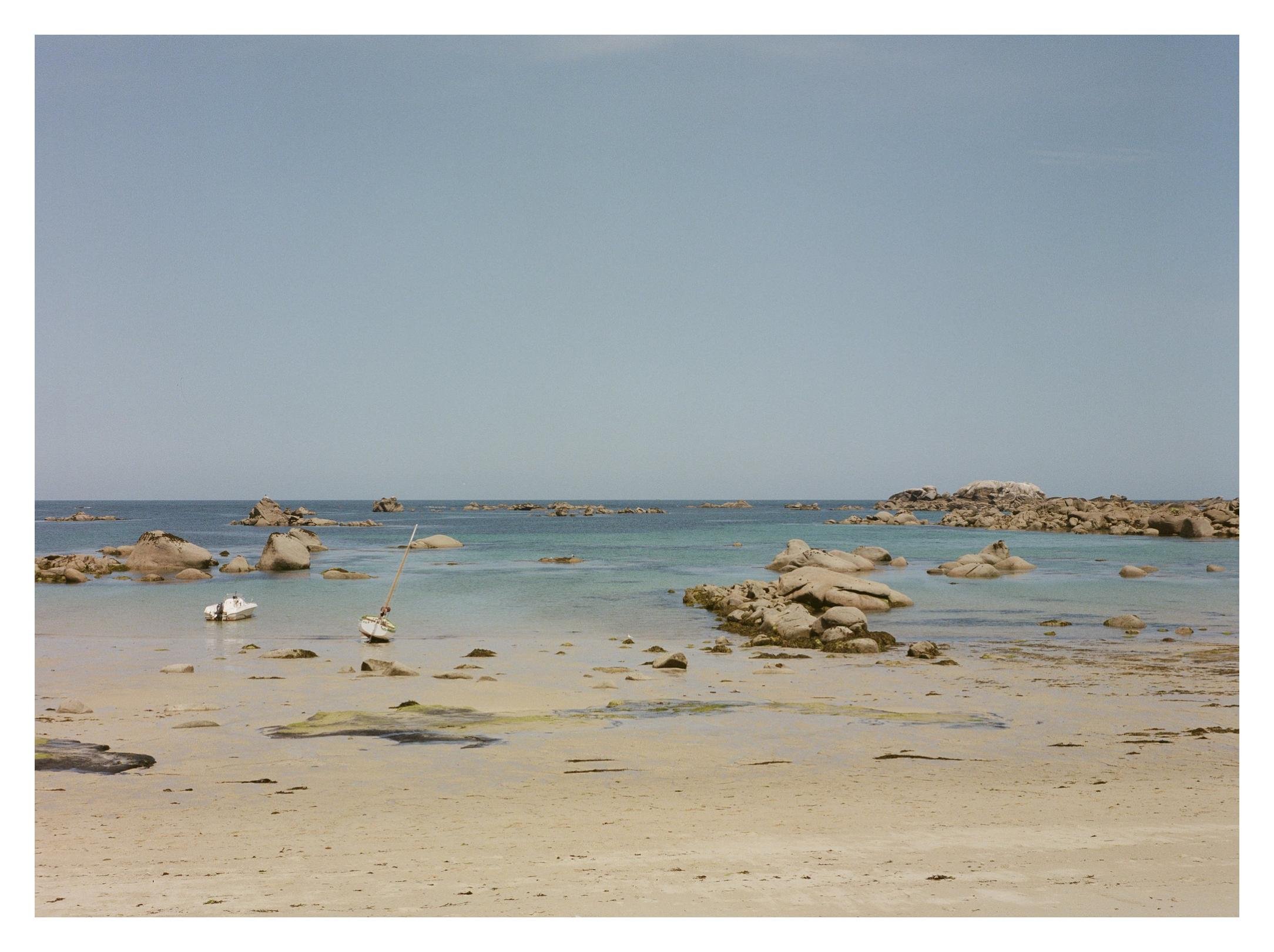 Sandy beach at low tide with shallow turquoise water, scattered seaweed and rocks, two small boats near shore, and a rocky outcrop along the horizon under a clear blue sky.