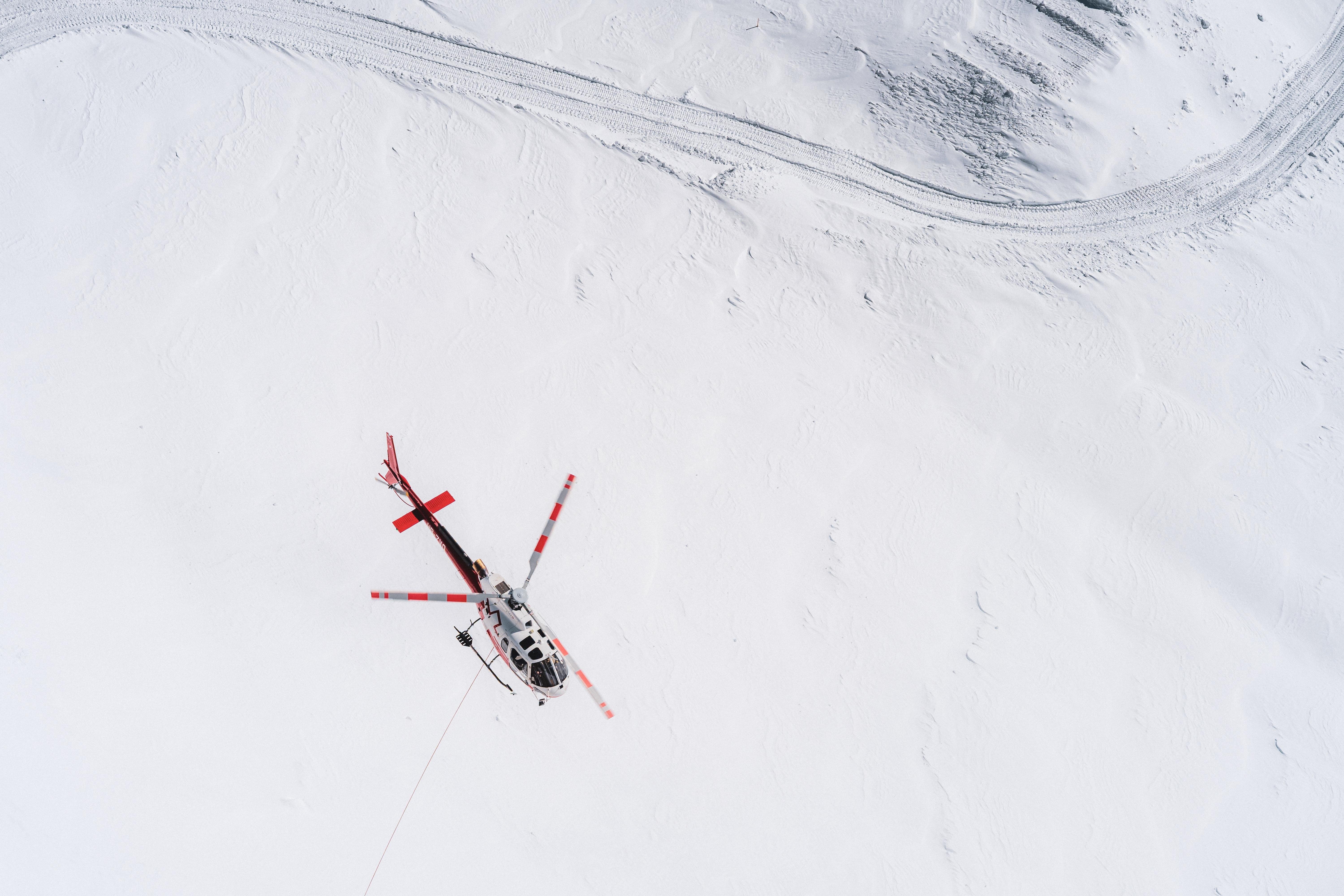 The image shows a helicopter flying over a snow-covered landscape. The helicopter has red accents and is seen from above, highlighting its spinning rotor blades. There are tracks in the snow, possibly from vehicles or machinery, visible in the top right corner of the image.