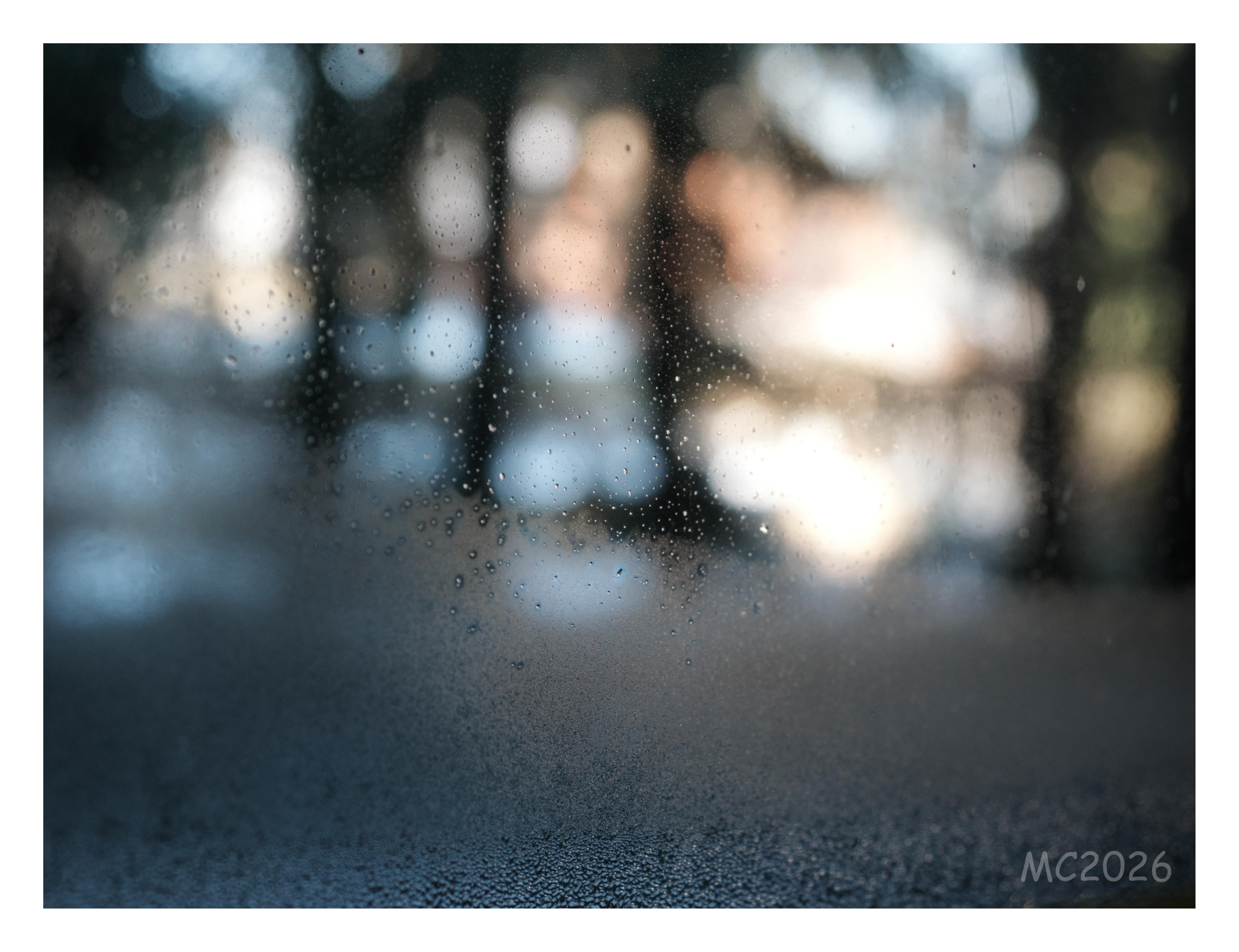 Drops of water on a window in sharp focus, with an out-of-focus background of trees and light-colored buildings; condensation covers the lower half of the glass.