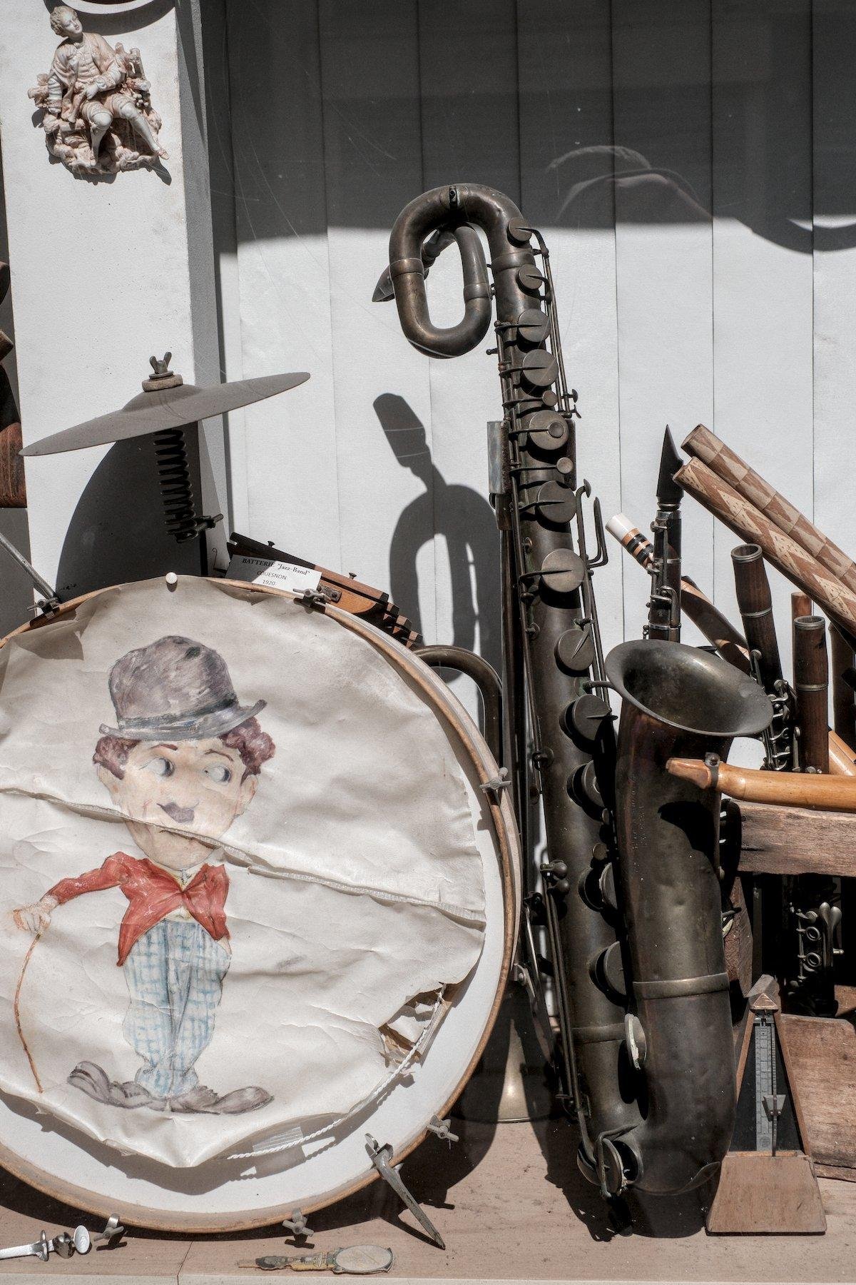 Photo verticale. Dans une vitrine, un très vieux saxophone noirci par le temps, au côté d’une grosse caisse de batterie décorée d’un Charly Chaplin en veste rouge, pantalon bleu, son célèbre chapeau melon et sa canne