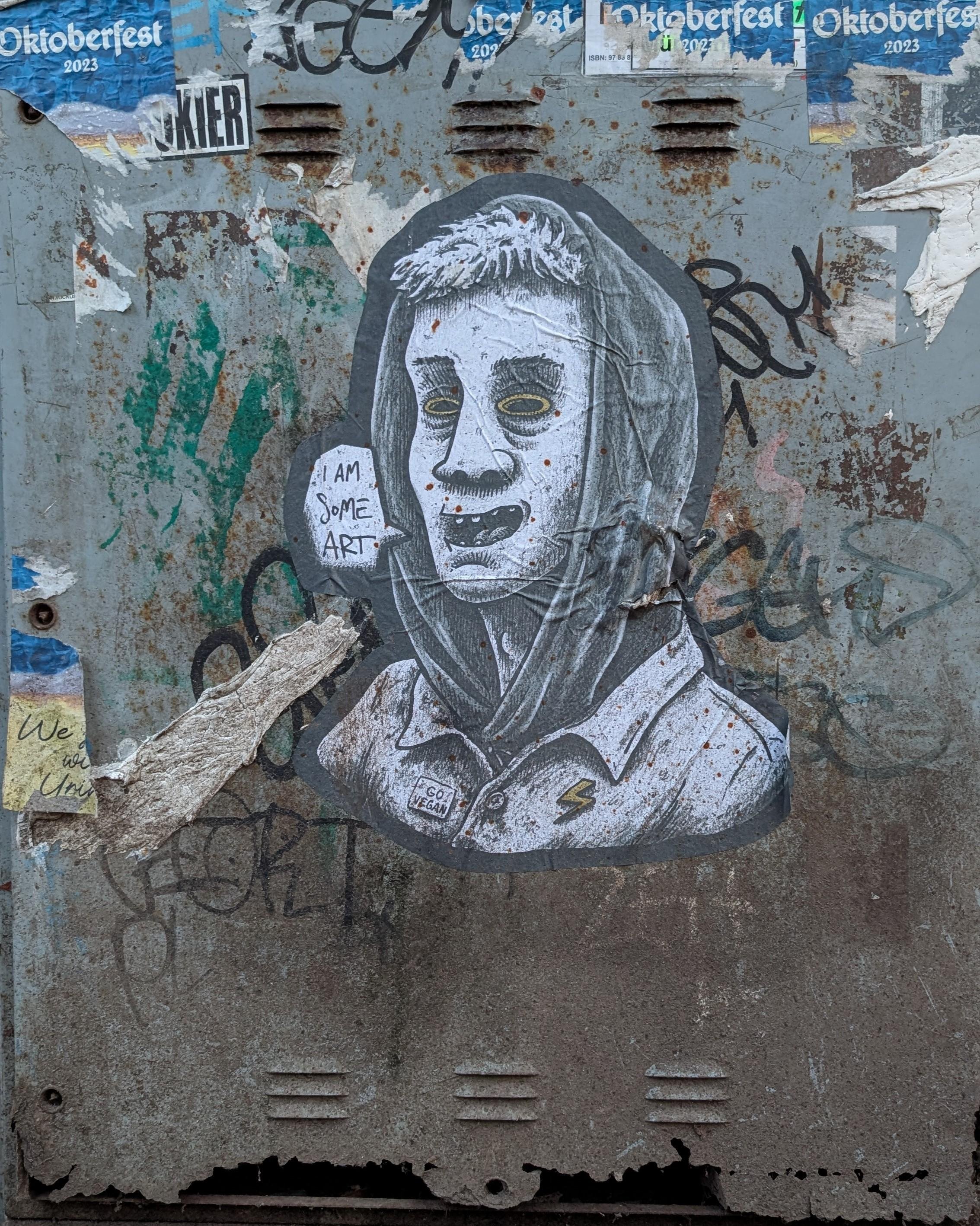 Pasted illustration of a person with exaggerated features, wearing a hoodie on a weathered surface. The surface is rusty and covered with graffiti, including remnant posters advertising "Oktoberfest 2023" and a peeled note with "We" visible. The figure has a textured, monochrome design with yellow eyes and an open mouth. Beside the figure is a speech bubble saying "I AM SOME ART." A badge reading "GO VEGAN" is on the collar.