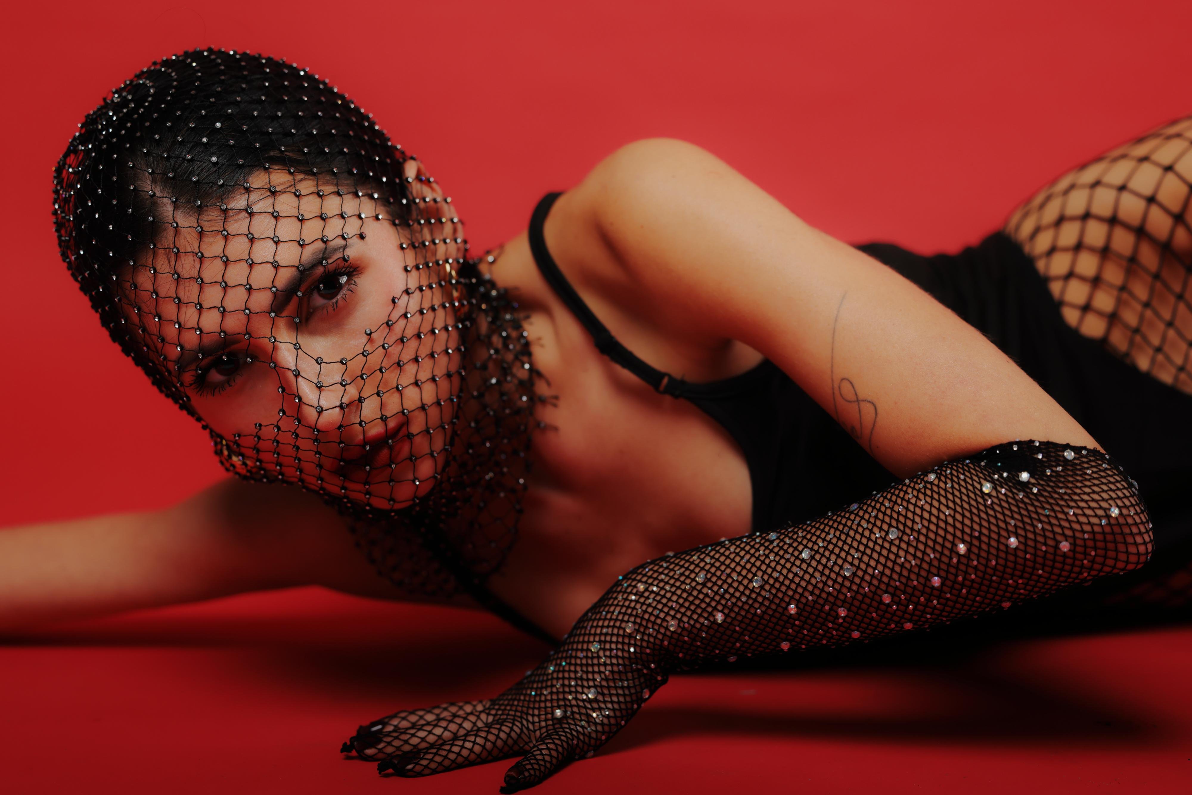 Woman in black attire lies on a red background, adorned with a netted headpiece and gloves embellished with pearls. Her left arm is extended forward, showcasing a minimalist tattoo near the wrist.