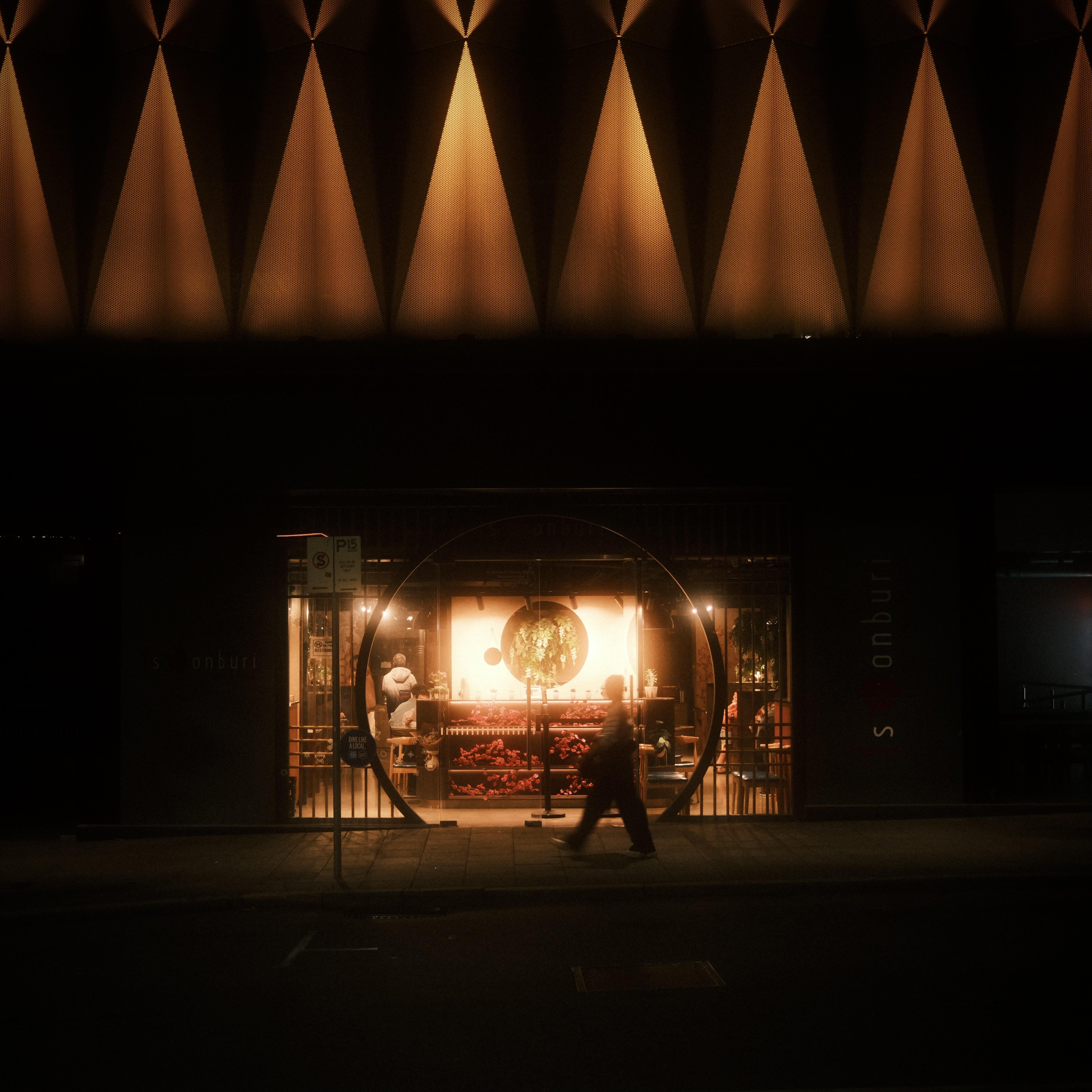 Night street view of a storefront with warm interior lighting, seen through a large circular window and a blurred pedestrian walking past in the foreground, with a row of triangular exterior lighted architectural features above.