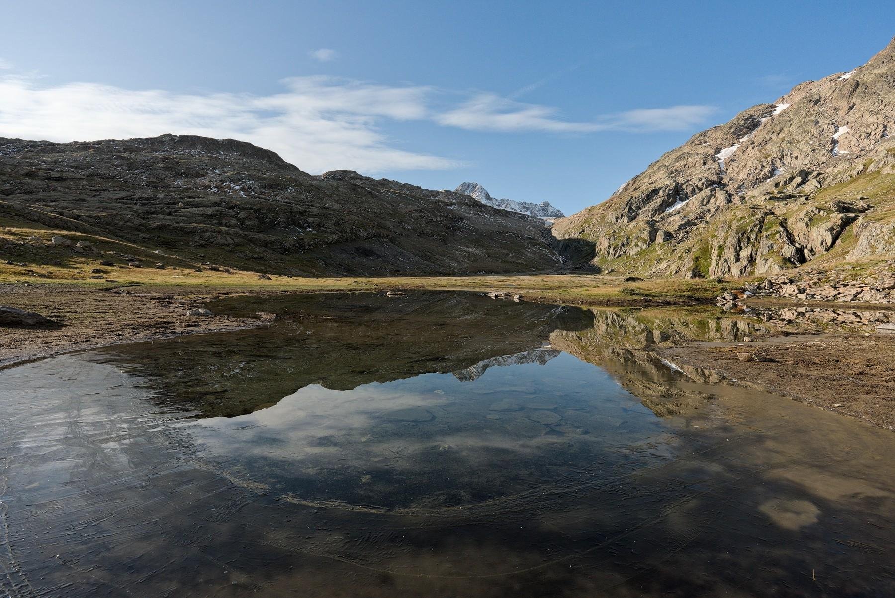 Cette image représente une scène naturelle tranquille avec un lac dont l'eau claire reflète parfaitement le paysage environnant. Le paysage est composé de montagnes escarpées, de terrains rocheux et de prairies herbeuses. Le ciel est généralement clair avec quelques nuages, et la scène dans son ensemble évoque un sentiment de calme et de beauté naturelle. Le premier plan montre le bord du lac légèrement gelé et l'arrière-plan est dominé par de grandes montagnes rocheuses.