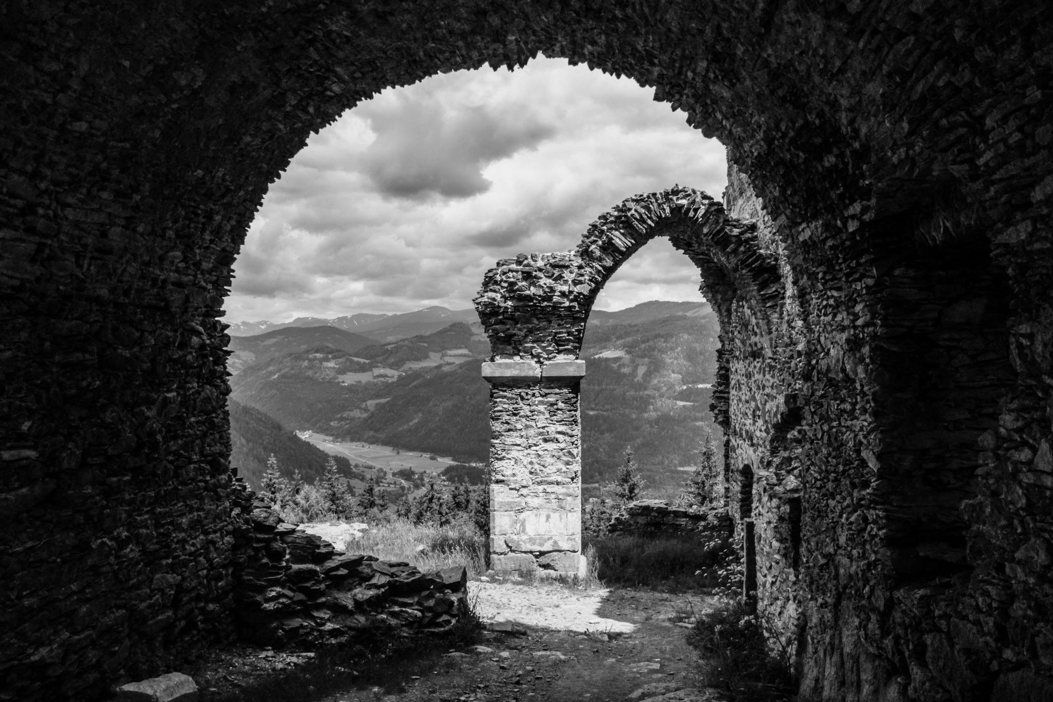 Black-and-white view from inside a stone ruin, framing a central stone pillar and partial arch, with a valley and layered mountains under cloudy sky in the background.