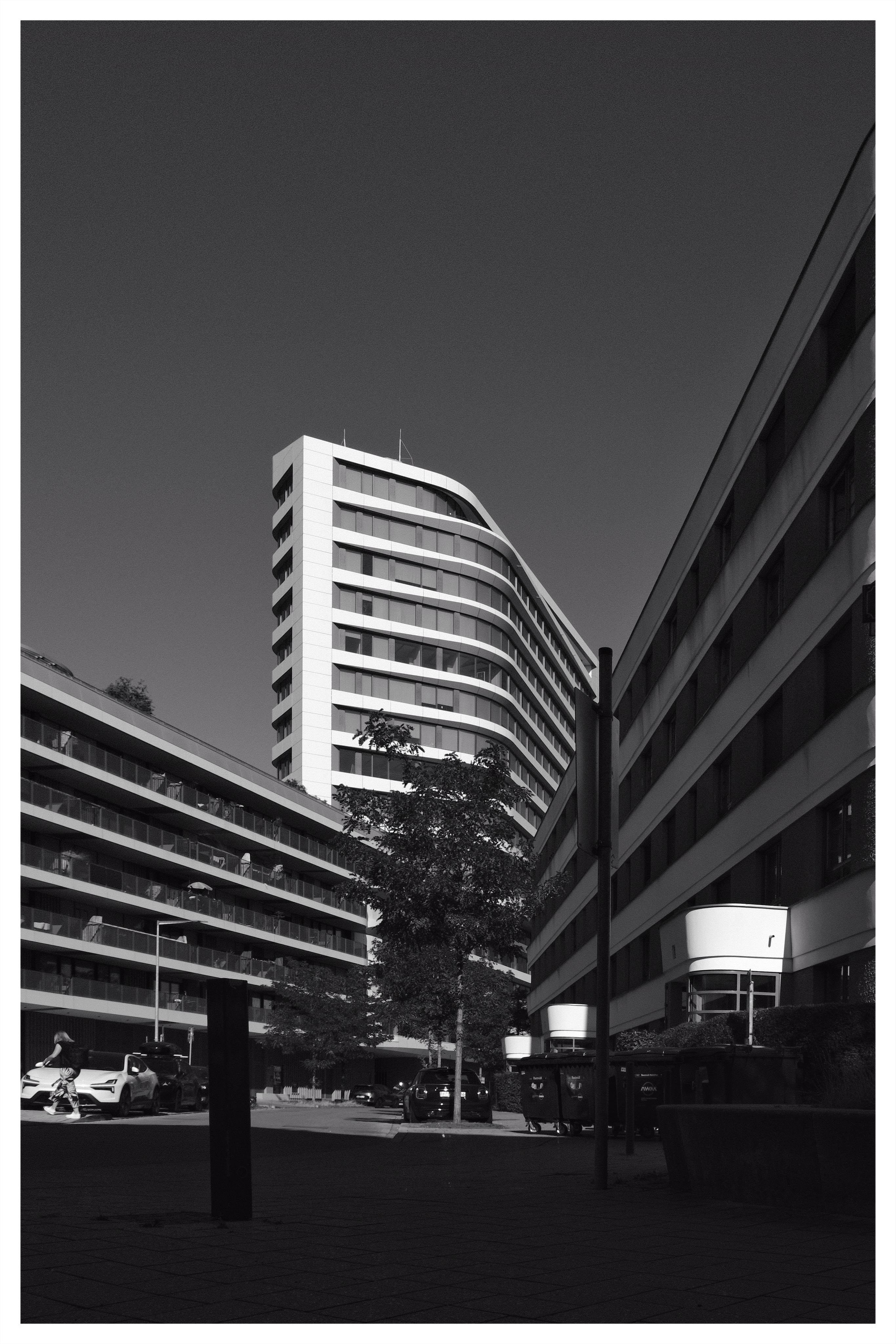 Black-and-white low-angle view of a modern curved high-rise centered between two long apartment blocks, with a small tree, streetlights, parked cars, and trash bins along a paved courtyard under a clear sky.