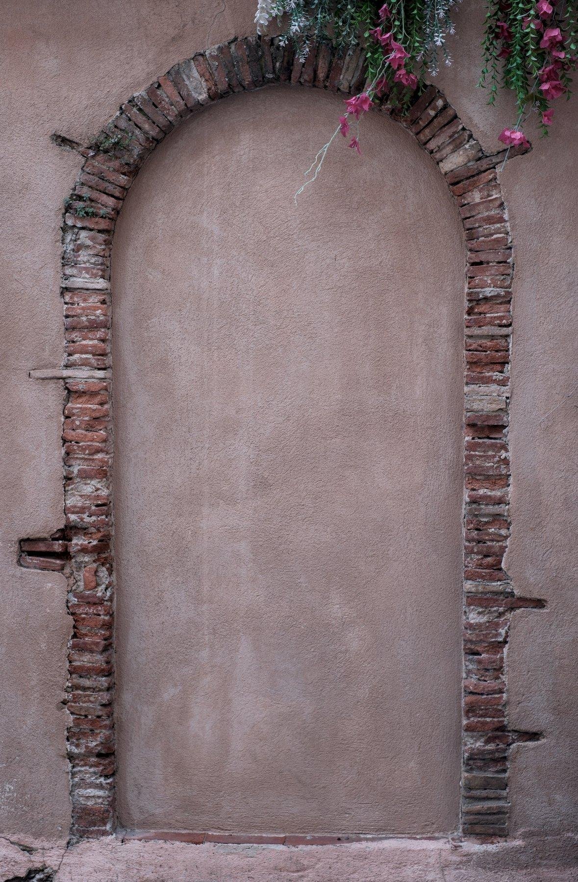 Vertical photo. In a uniform(ish) wall, an arch constructed with a stack of tiles of the same size, except for 7 of them which are twice the length, the whole drawing a past door
