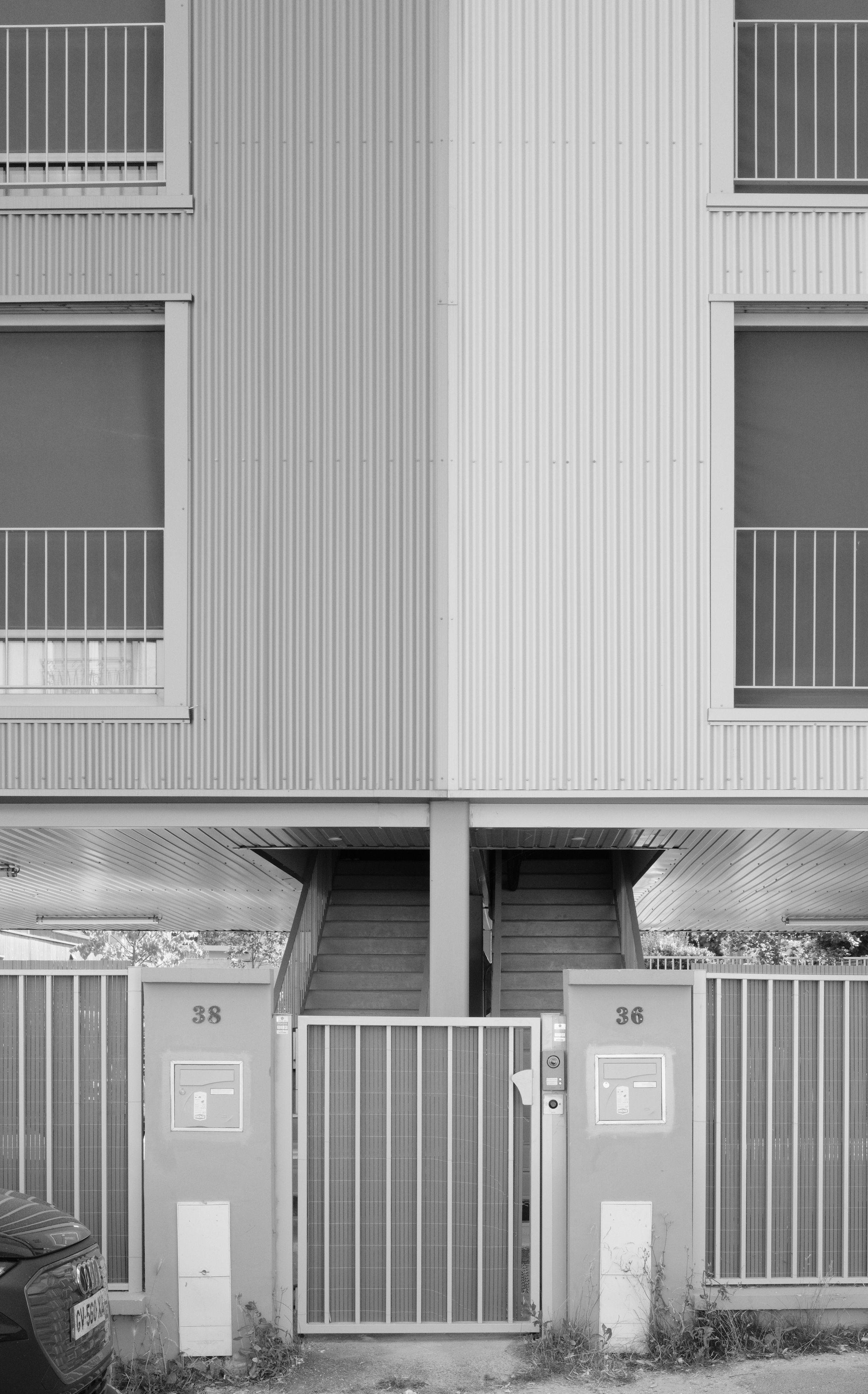 Black and white photo of a stairway shared by two buildings. Each building is of different colours, one white, one gray. Some fence protect them and a low fence door accessing the stairway is visible at the center, with its concrete columns