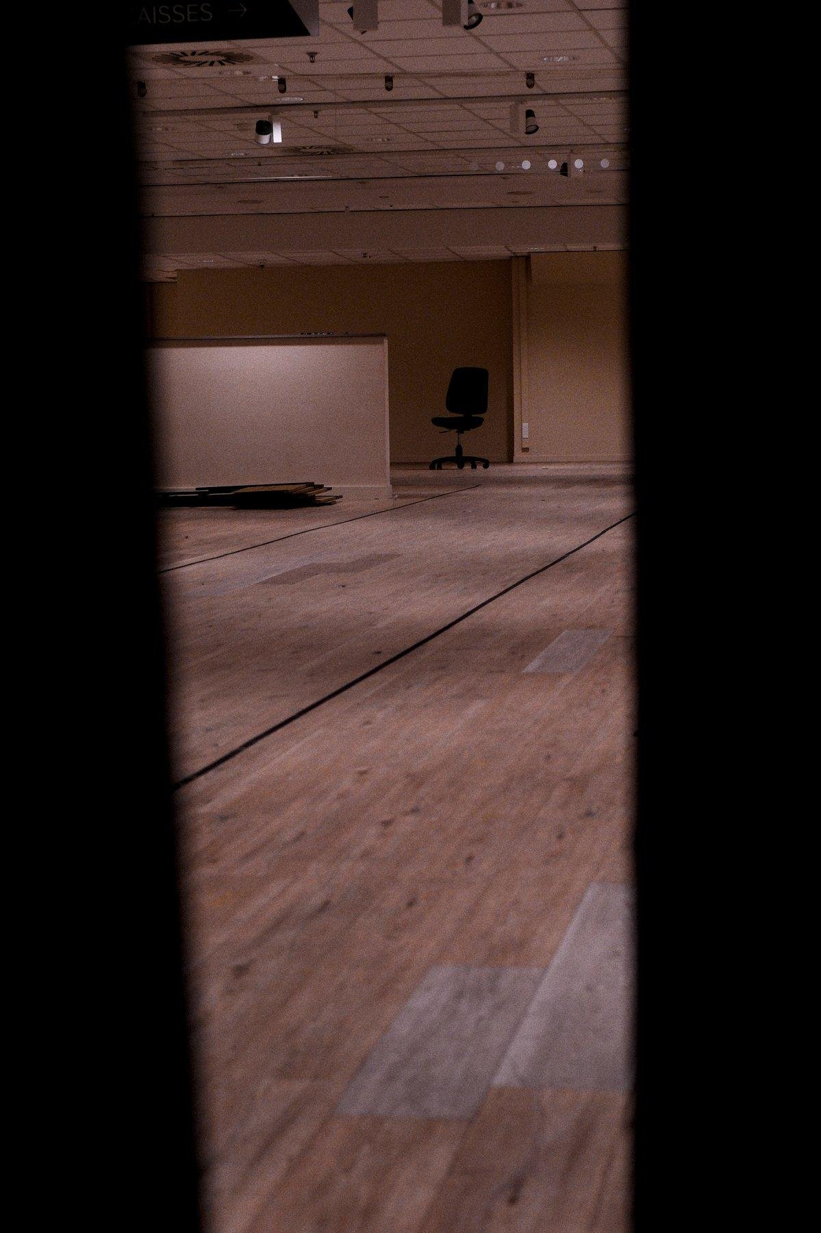 Photo verticale. Au premier plan, deux bandes noires de part et d’autre de l’image, comme si on regardait entre deux plaques. On voit un vaste espace vide avec un plancher sur lequel sont dessinées des marques noires délimitant des rectangles. Au fond, une chaise de bureau noire trône au milieu de nul part. Le tout est éclairé dans des teintes chaudes