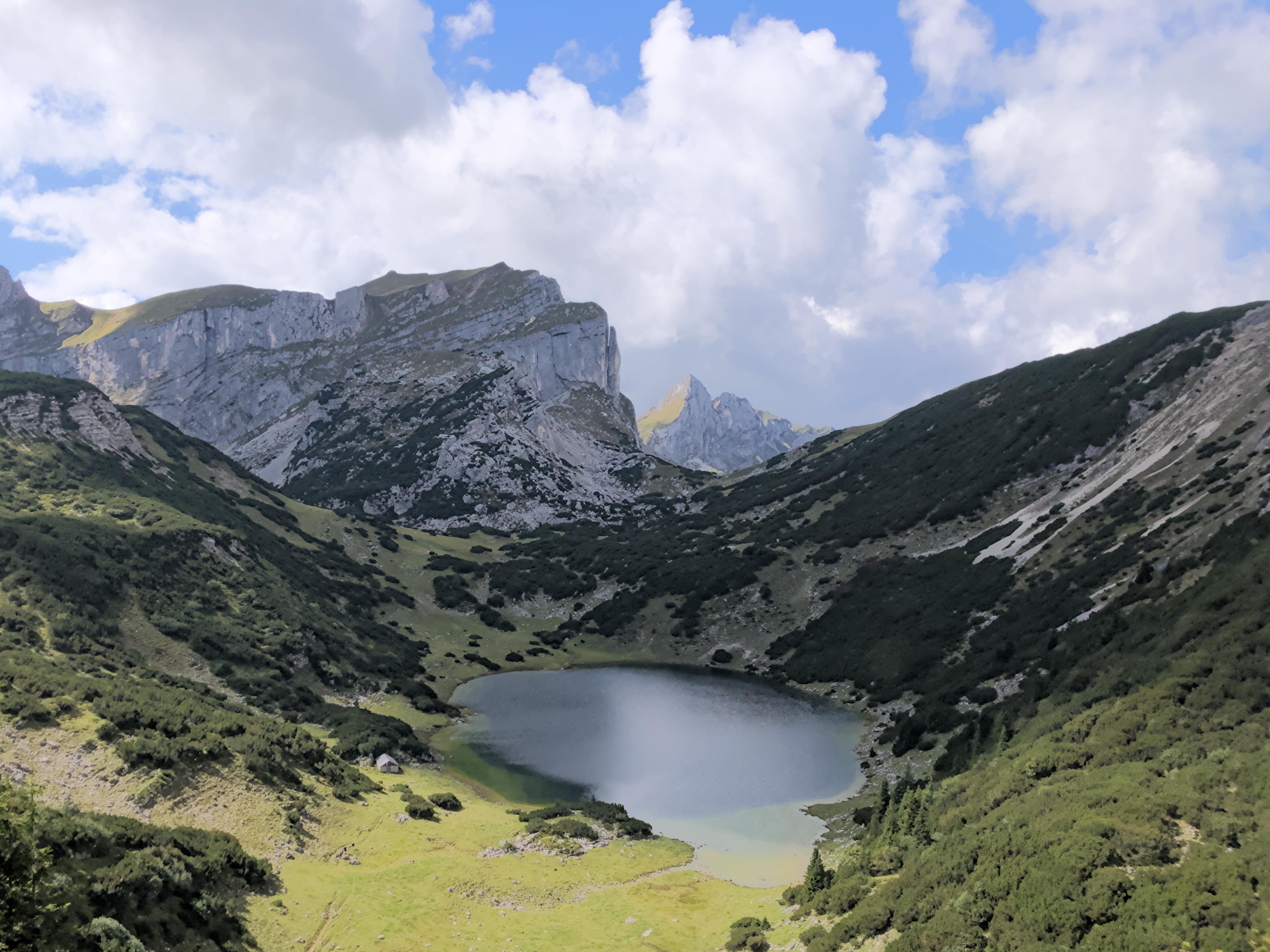 This image features a scenic landscape of a mountain range with a small lake or pond nestled in a lush, green valley. The mountains are rocky and have some vegetation, and the sky is partly cloudy with patches of blue visible. The area looks serene and natural.