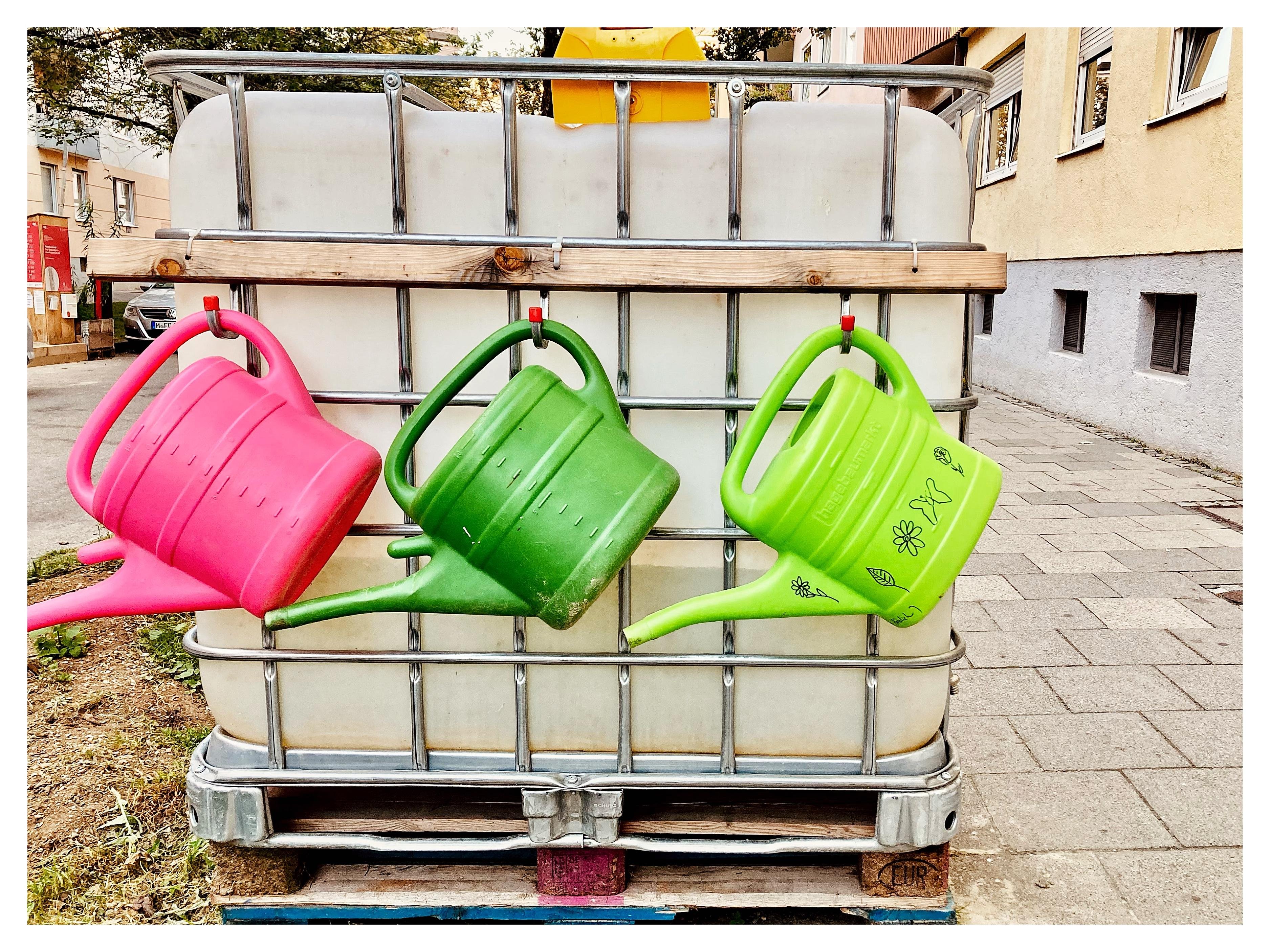 Three plastic watering cans—pink, dark green, and light green—hang from a wooden slat on the metal cage of a large translucent water tank on a pallet beside a sidewalk and building wall.