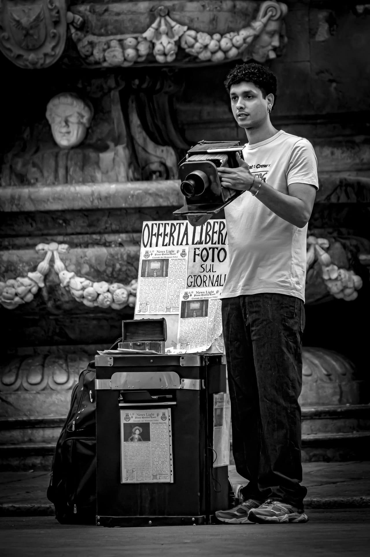 Black-and-white street scene of a young man standing beside a cart, holding a large instant camera. A sign on the cart reads “OFFERTA LIBERA” and “FOTO SUL GIORNALI,” with sample newspaper-style prints displayed; an ornate stone fountain fills the background.