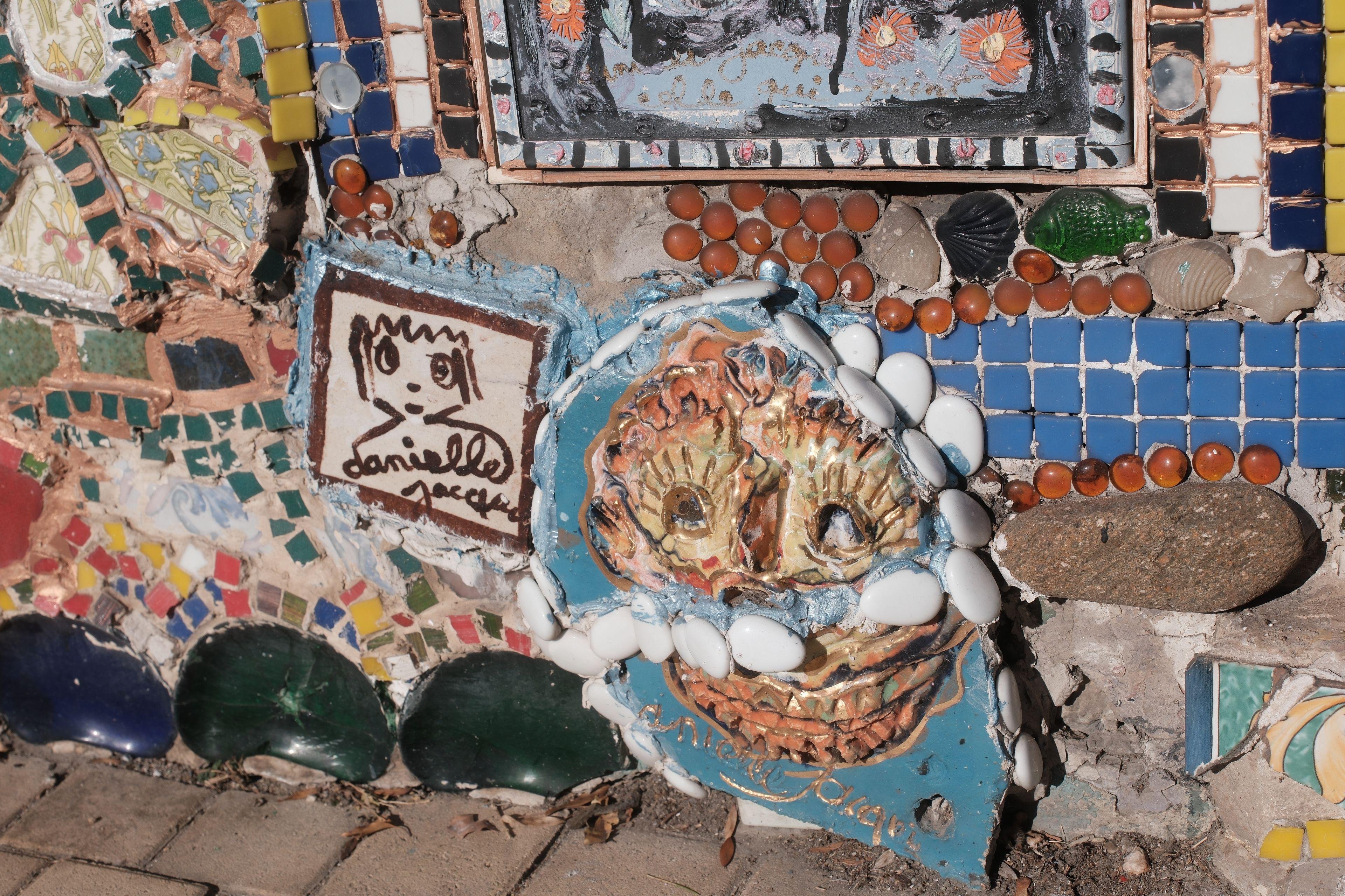 Photo format paysage d’un bout mur totalement décoré de céramiques en tout genre, mais la photo se concentre sur une espèce de soleil aux dents acérées et au regard fou, tout en teintes orangées sur fond de bleu turquoise