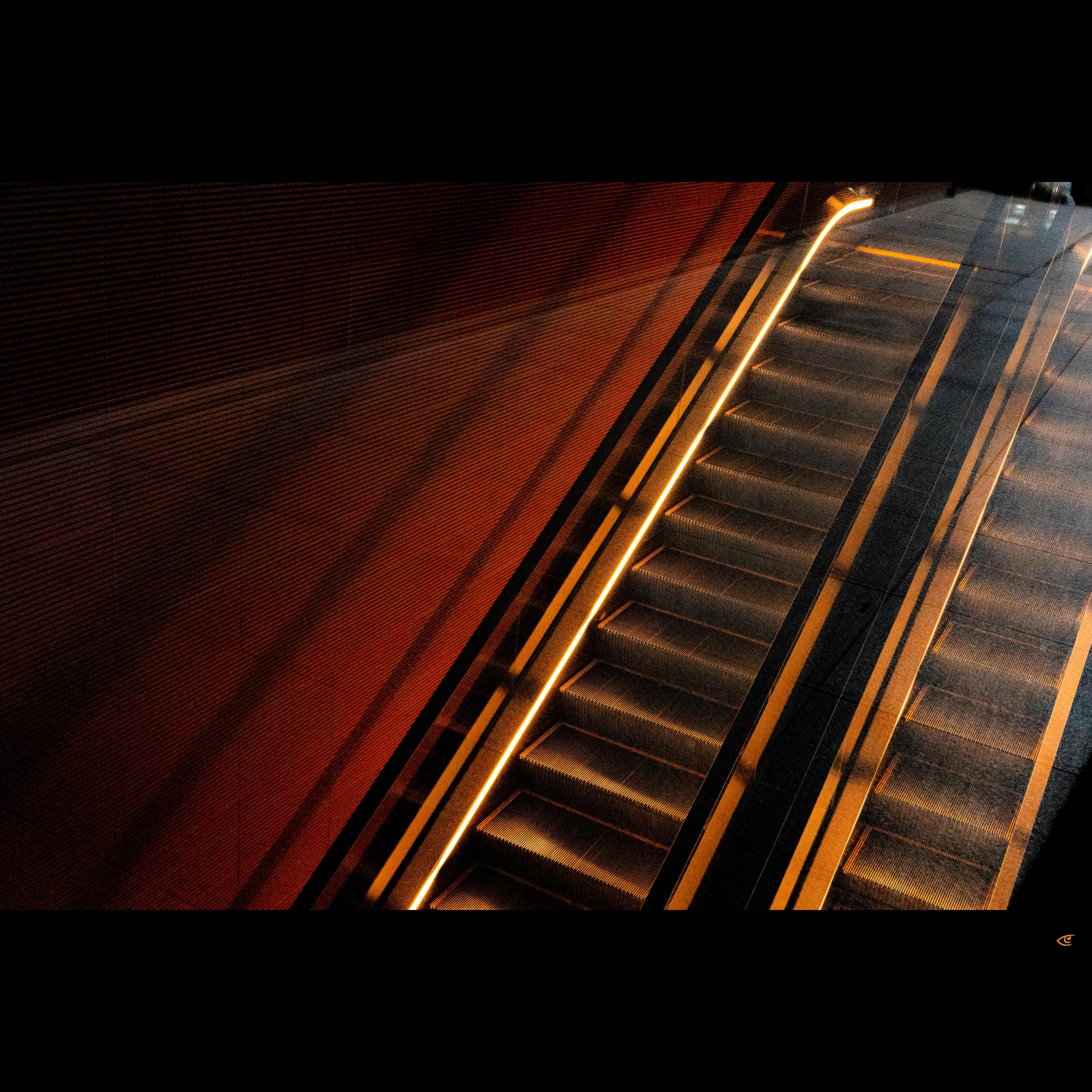 Diagonal overhead view of an escalator with illuminated handrails running upward beside a textured orange wall, with a second escalator partially visible on the right.