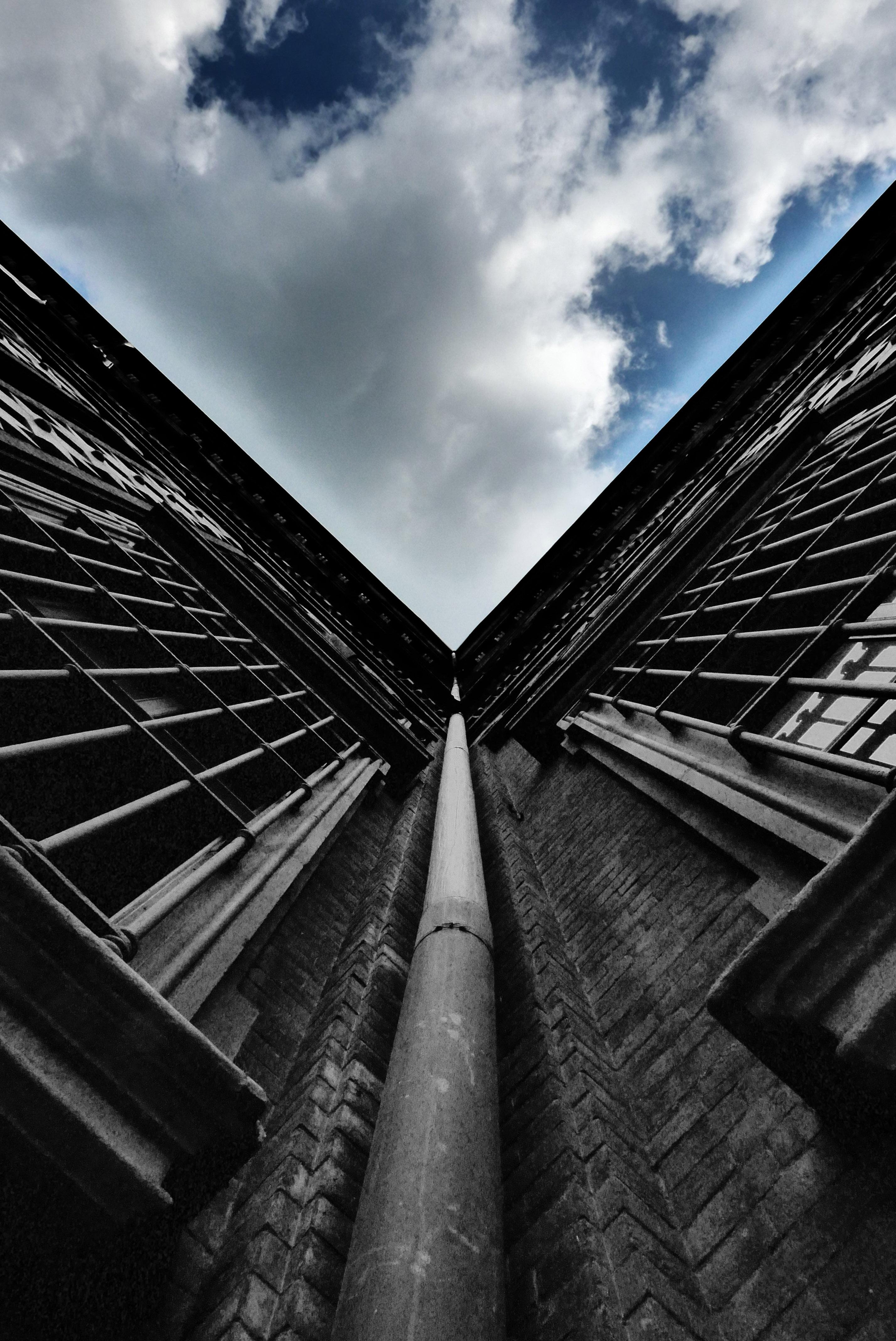 This image shows an upward perspective of a building, capturing the corner where two sides meet. It features brick walls with large windows and a central downspout. The sky above is partly cloudy, providing a contrasting backdrop. The angle emphasizes the height and architectural details of the structure.