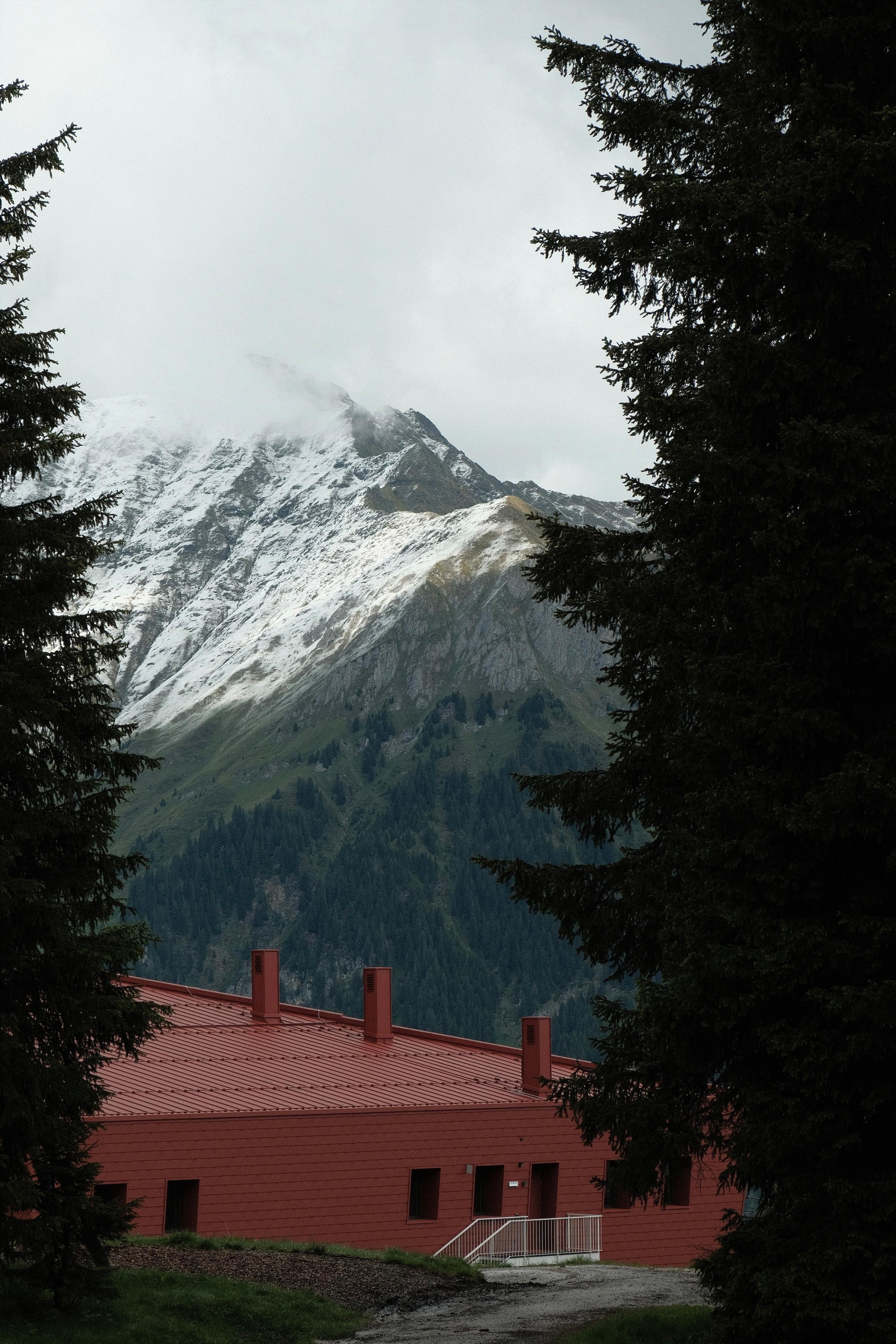 A Mountain View framed by two dark trees on the left and right side. In the lower foreground a red modern minimalistic building contrast the natural landscape of a mountain with a snow cap in the background. A bright white staircase at the side of the red building contrasts the darker foreground and resonates with the white snow cap in the upper part. The sky is cloudy.