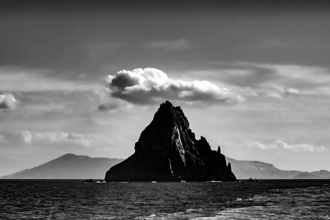 Black-and-white seascape with a jagged rocky sea stack centered in the water, low distant mountains on the horizon, and layered clouds across the sky.