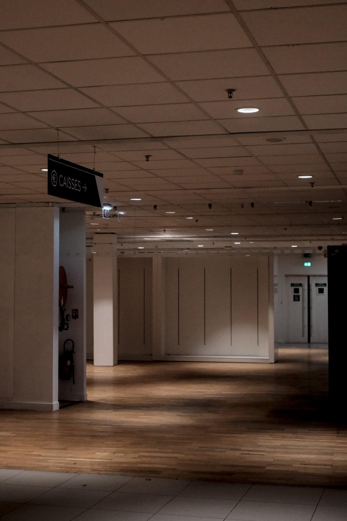 Photo verticale. Espace sans commerce, sans client, d’une galerie marchande. Un panneau suspendu indique la direction des caisses désormais disparues, et on voit une porte de secours au bout de la direction indiquée