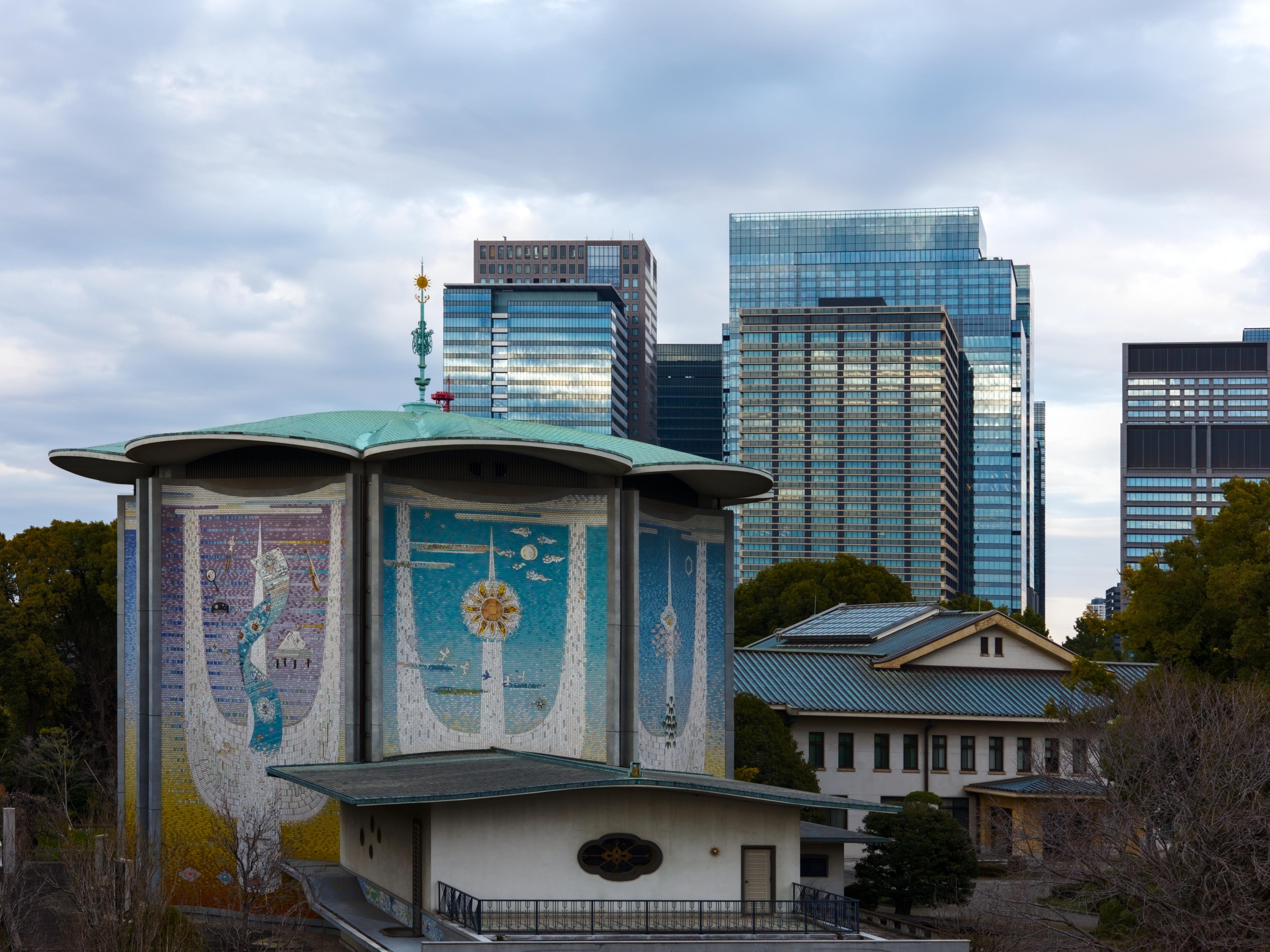 A traditional, colorful building from the Tokyo Imperial Palace, standing in front of glass and steel modern buildings, with a gray, cloudy sky.